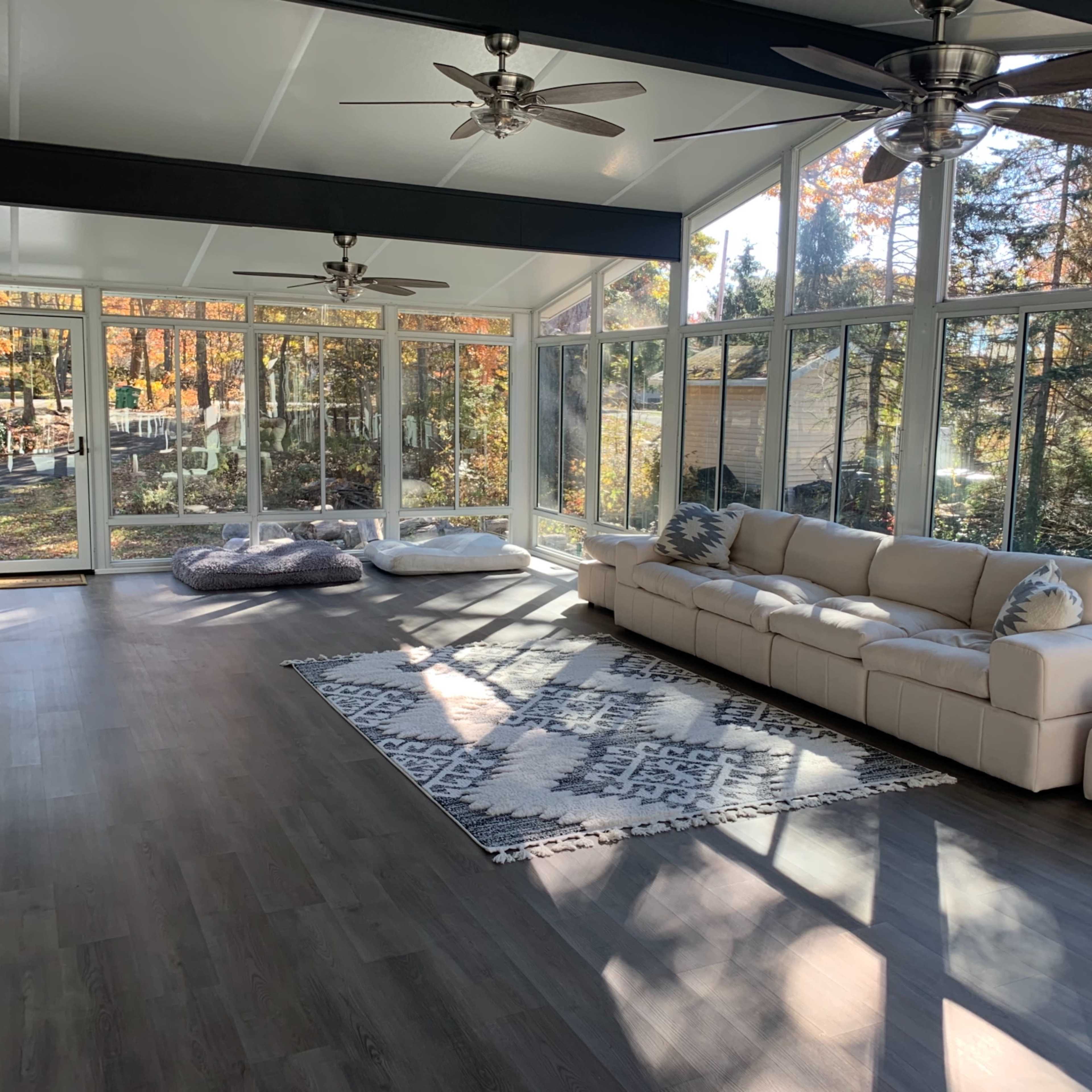 A bright sunroom with large windows, a sectional sofa, a patterned rug, and two ceiling fans.