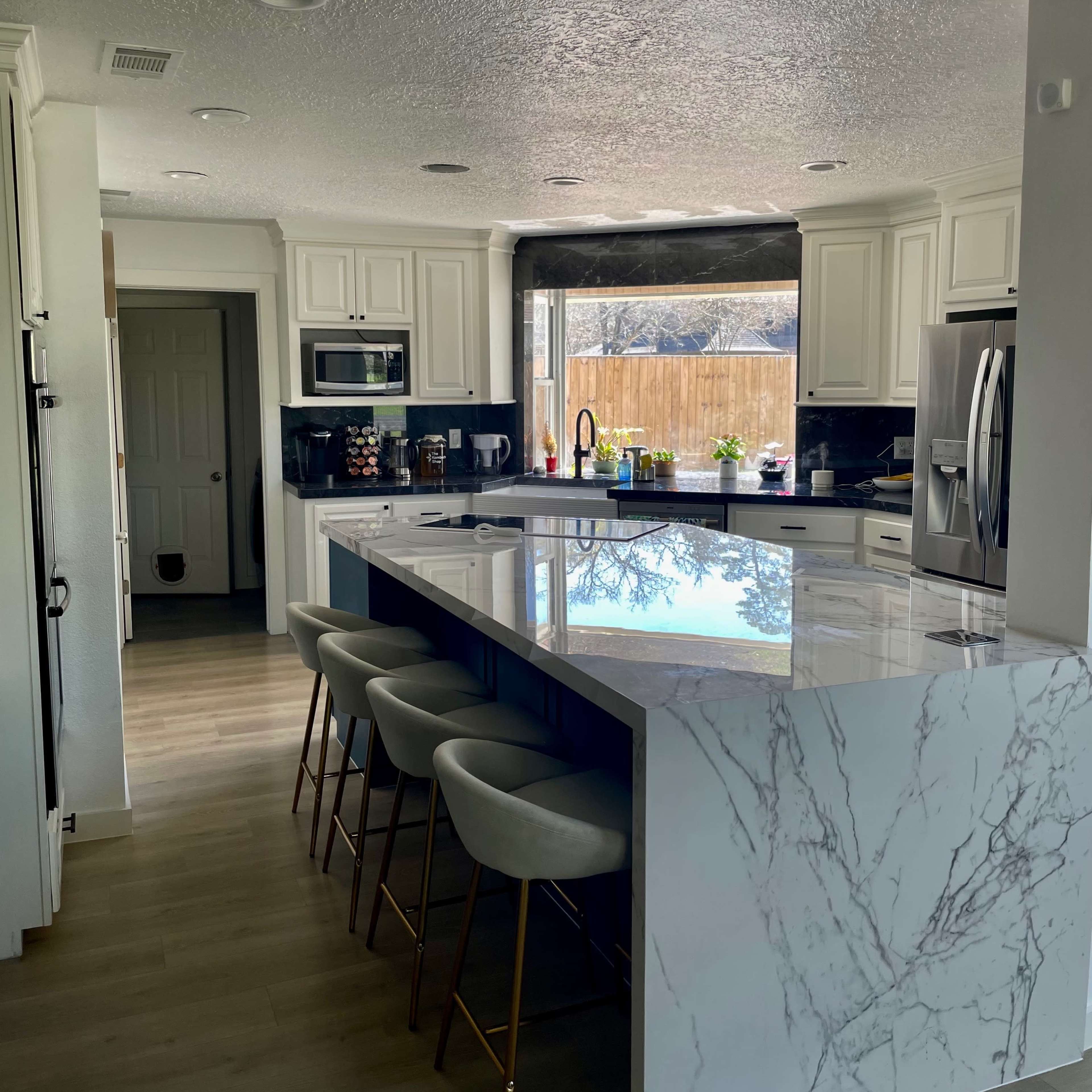 A modern kitchen featuring white cabinetry, a large marble island with seating, and a view of a backyard through a large window.
