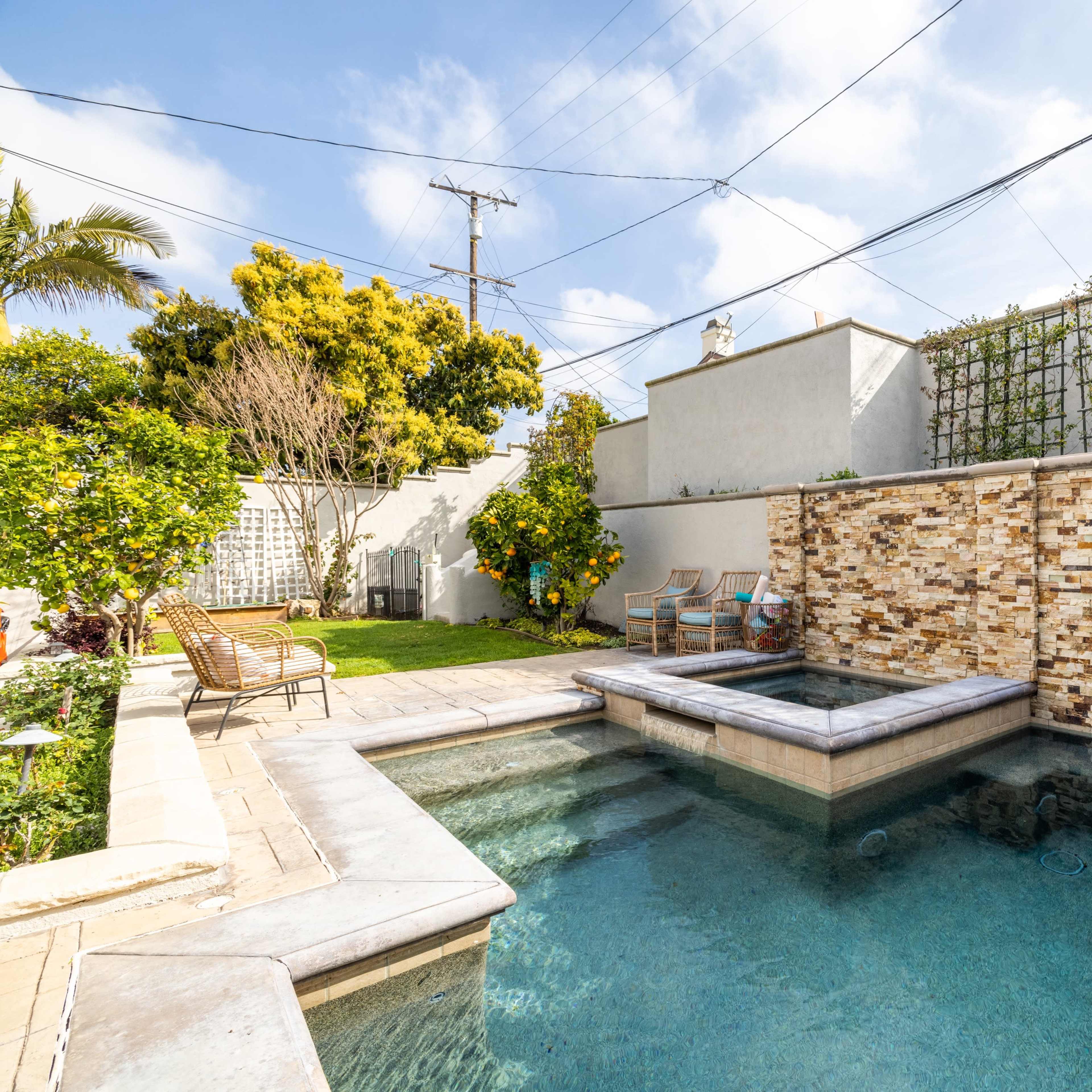 A backyard pool area featuring a stone wall, a spa, and several lounge chairs surrounded by greenery.