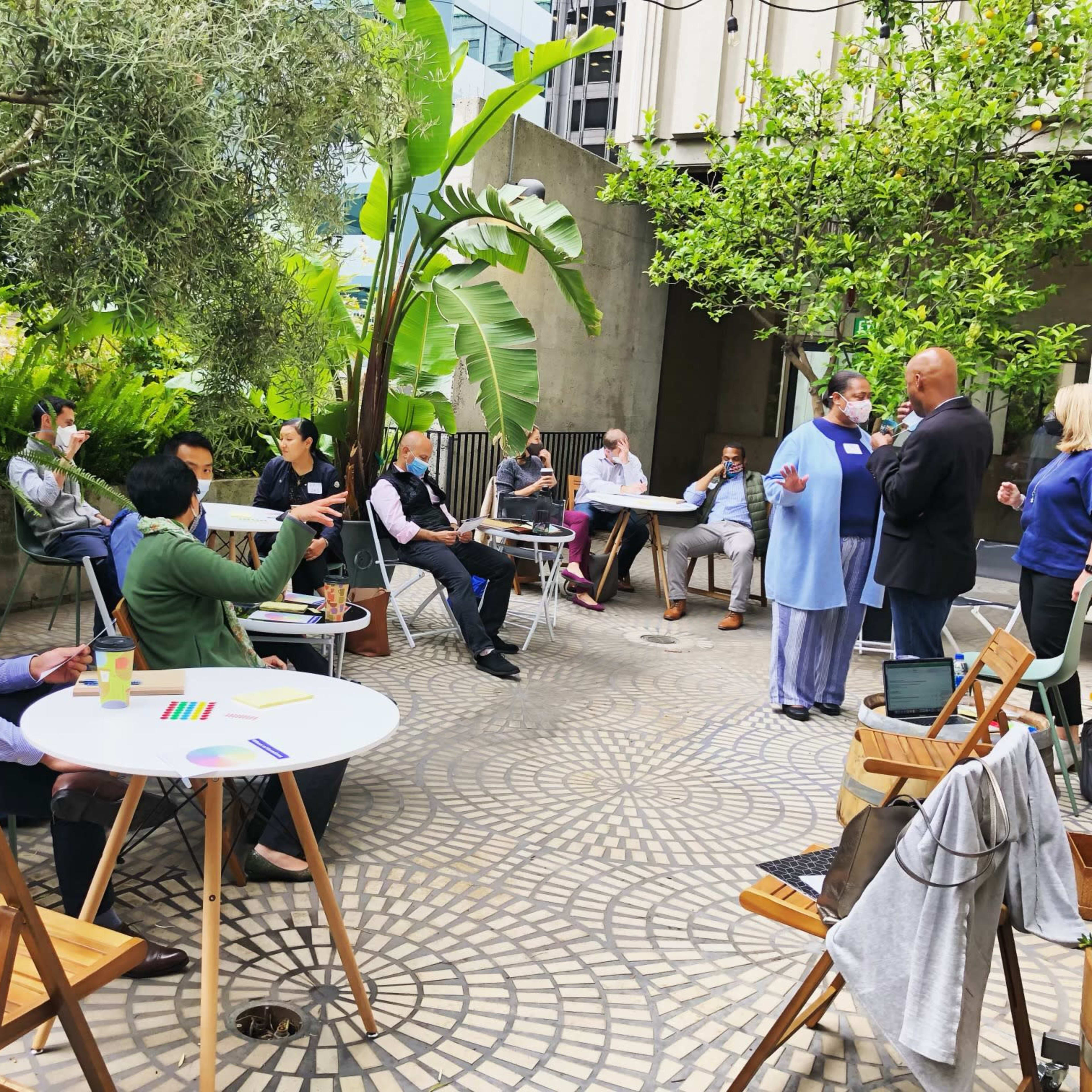 A group of people is engaged in discussions around tables in a landscaped outdoor setting.