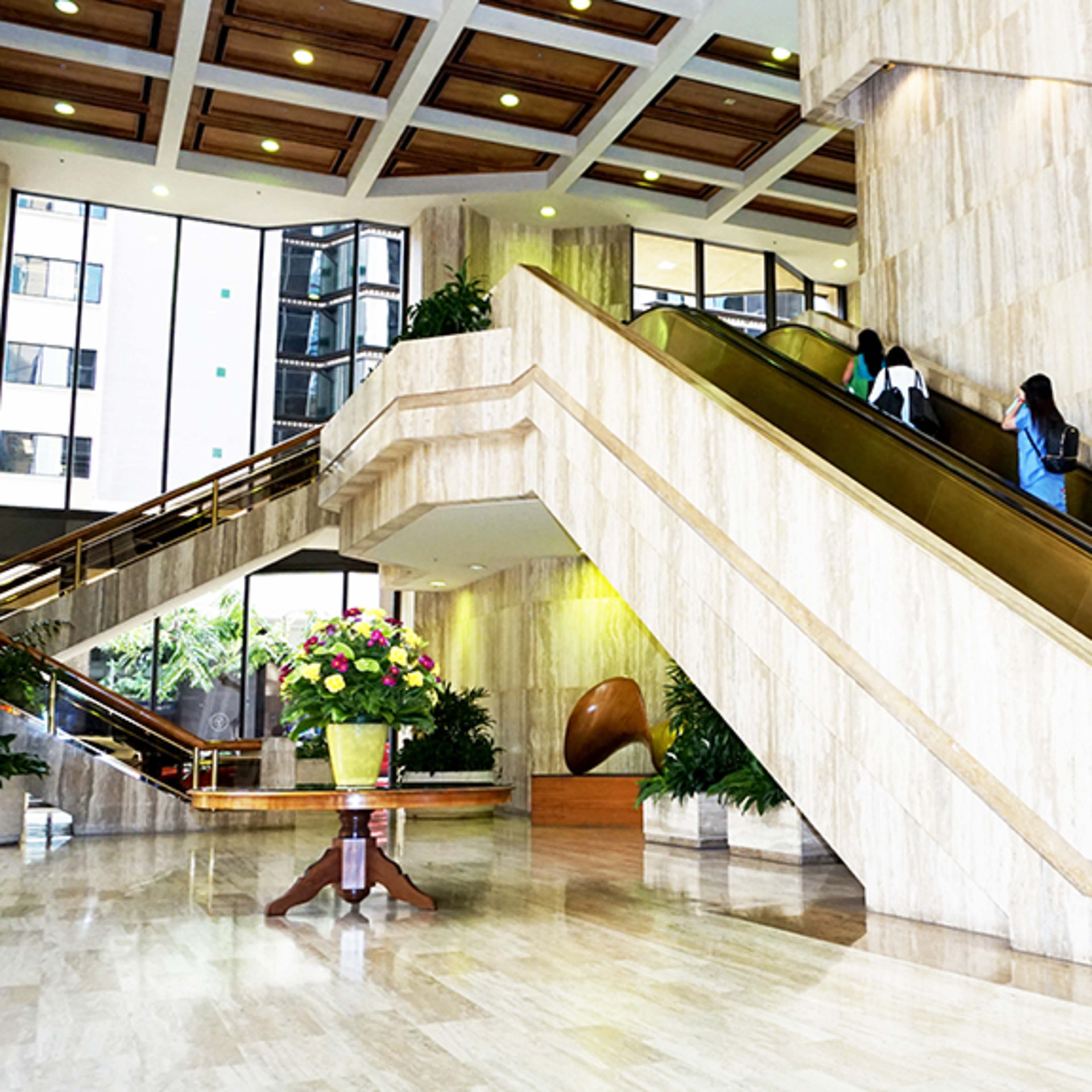 A spacious lobby with marble walls and two escalators leading to an upper level, flanked by large plants and a floral arrangement in the foreground.