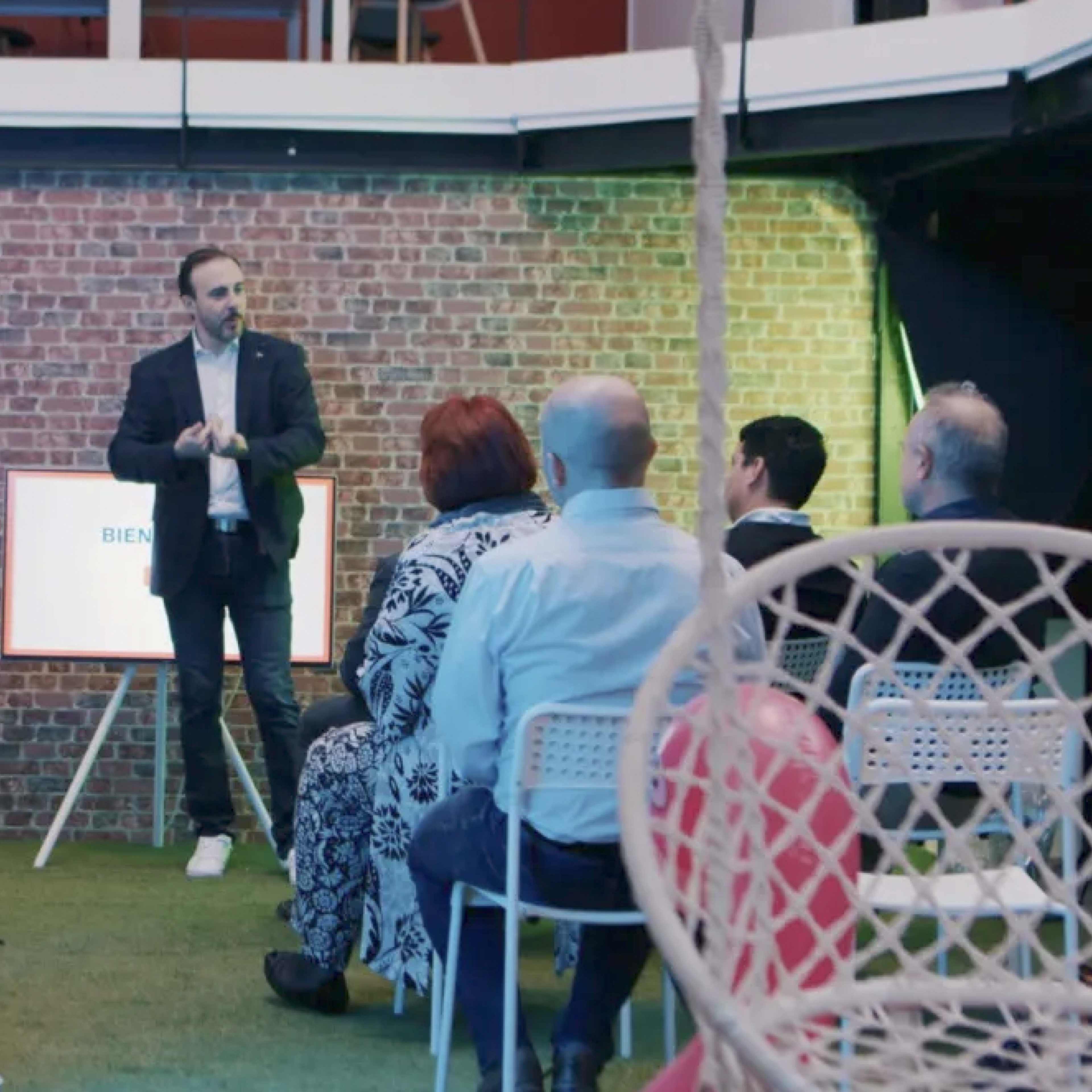 A speaker stands in front of a seated audience in a space decorated with a brick wall and greenery.