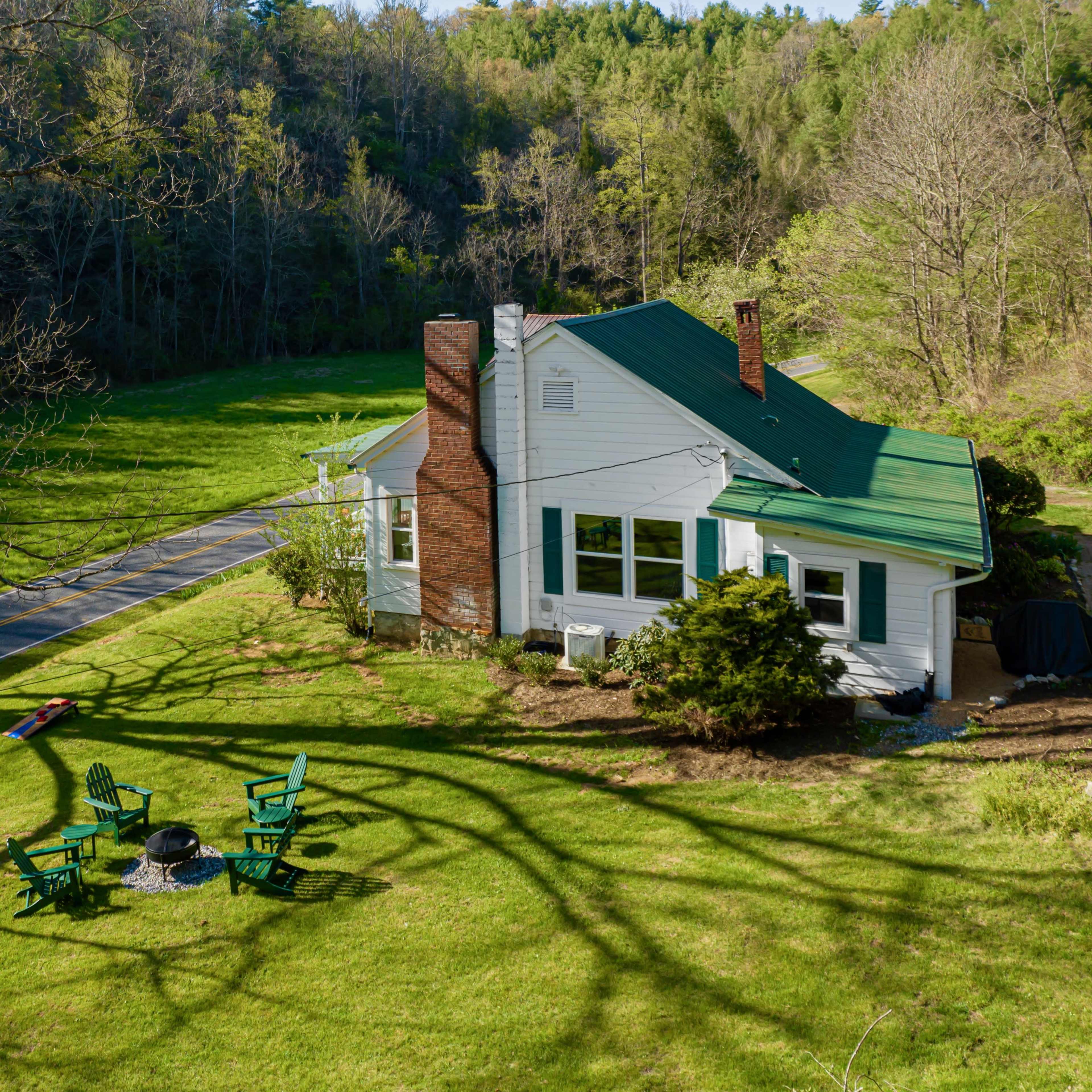 A house with a green roof sits next to a winding road, surrounded by a lawn featuring a fire pit and seating area.