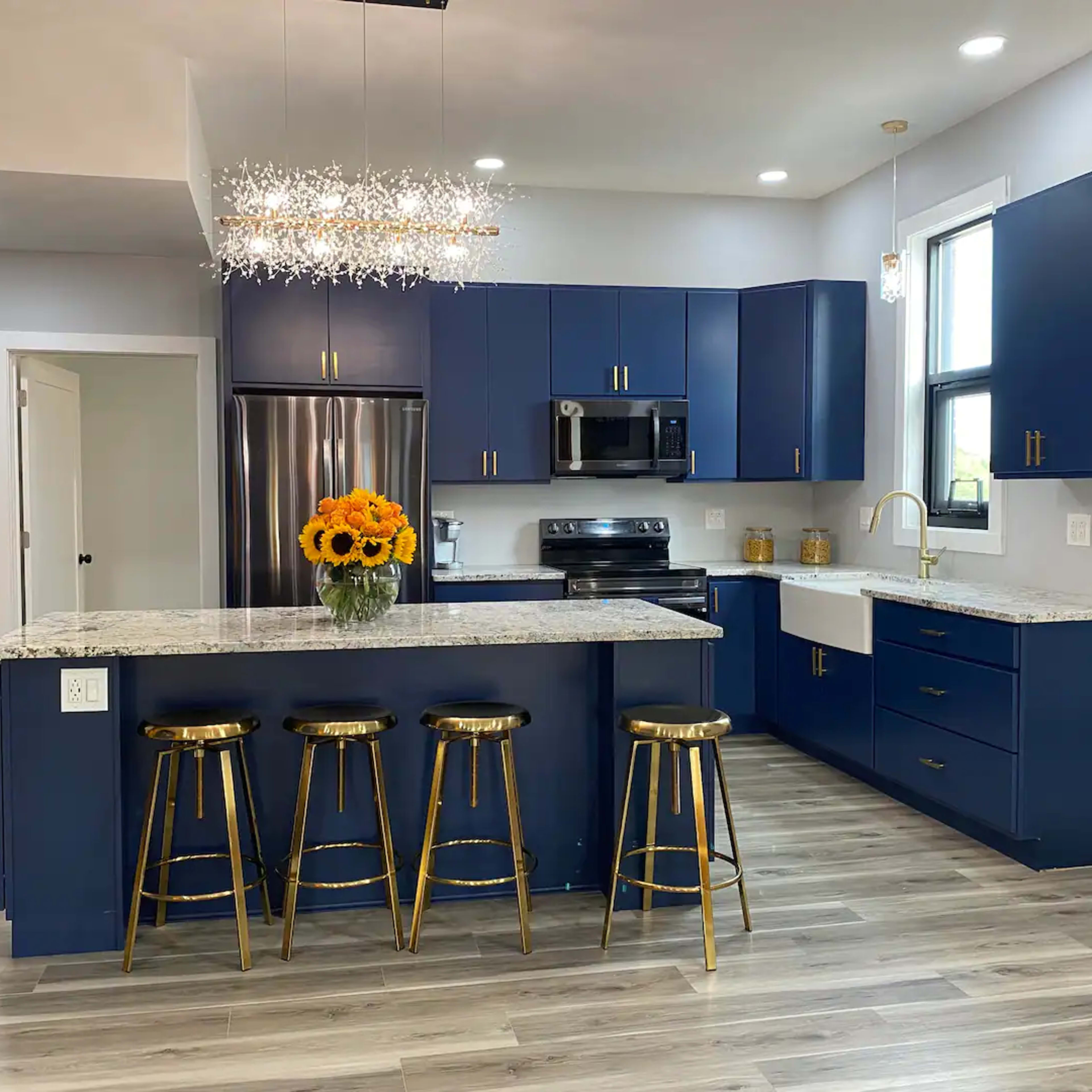 A modern kitchen with navy blue cabinets, a granite countertop, and a chandelier above an island with four gold bar stools.