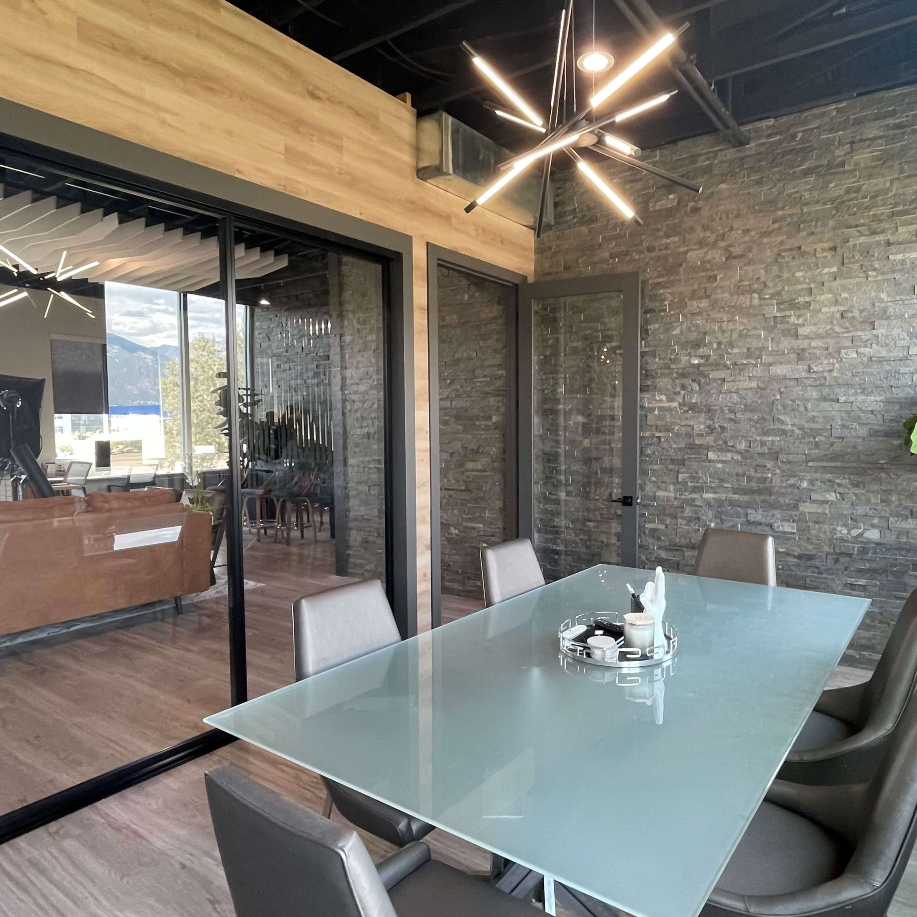 A modern conference room features a glass table surrounded by chairs, with a stone accent wall and a large window overlooking an adjacent space.