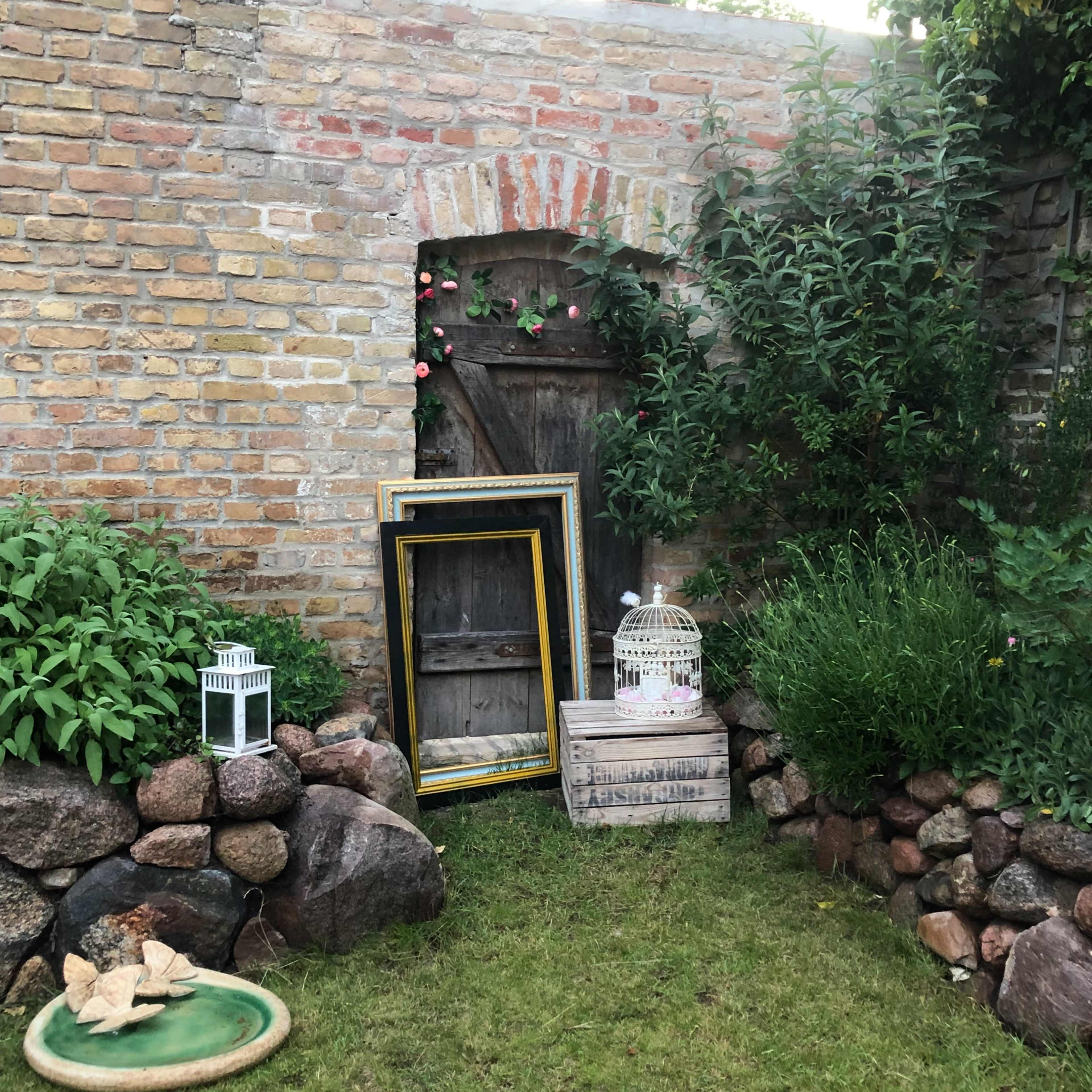 An old brick wall features a wooden door surrounded by greenery and decorative items, including empty frames and lanterns, set against a grassy area.