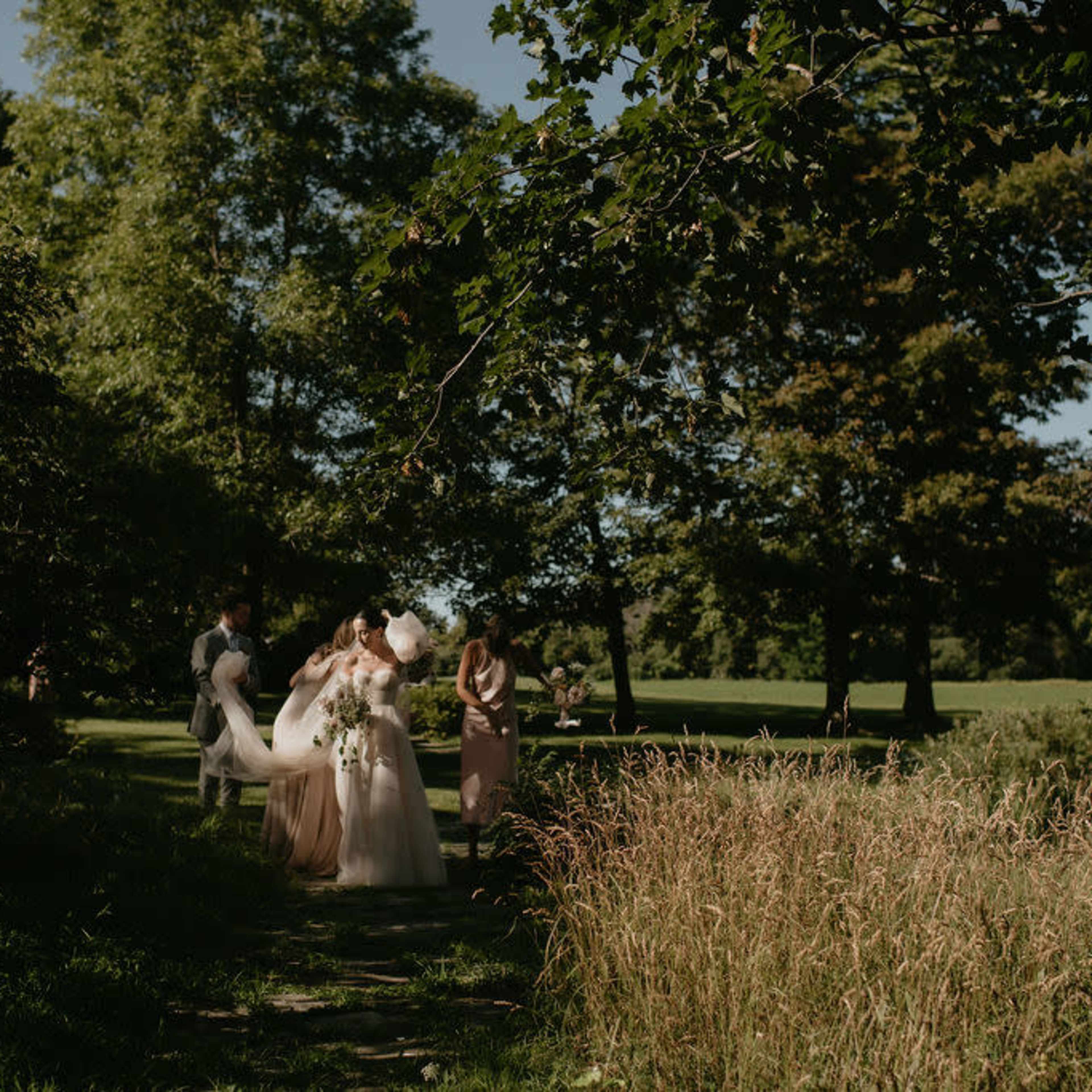 A bride in a white dress stands on a path surrounded by greenery, accompanied by four other individuals.