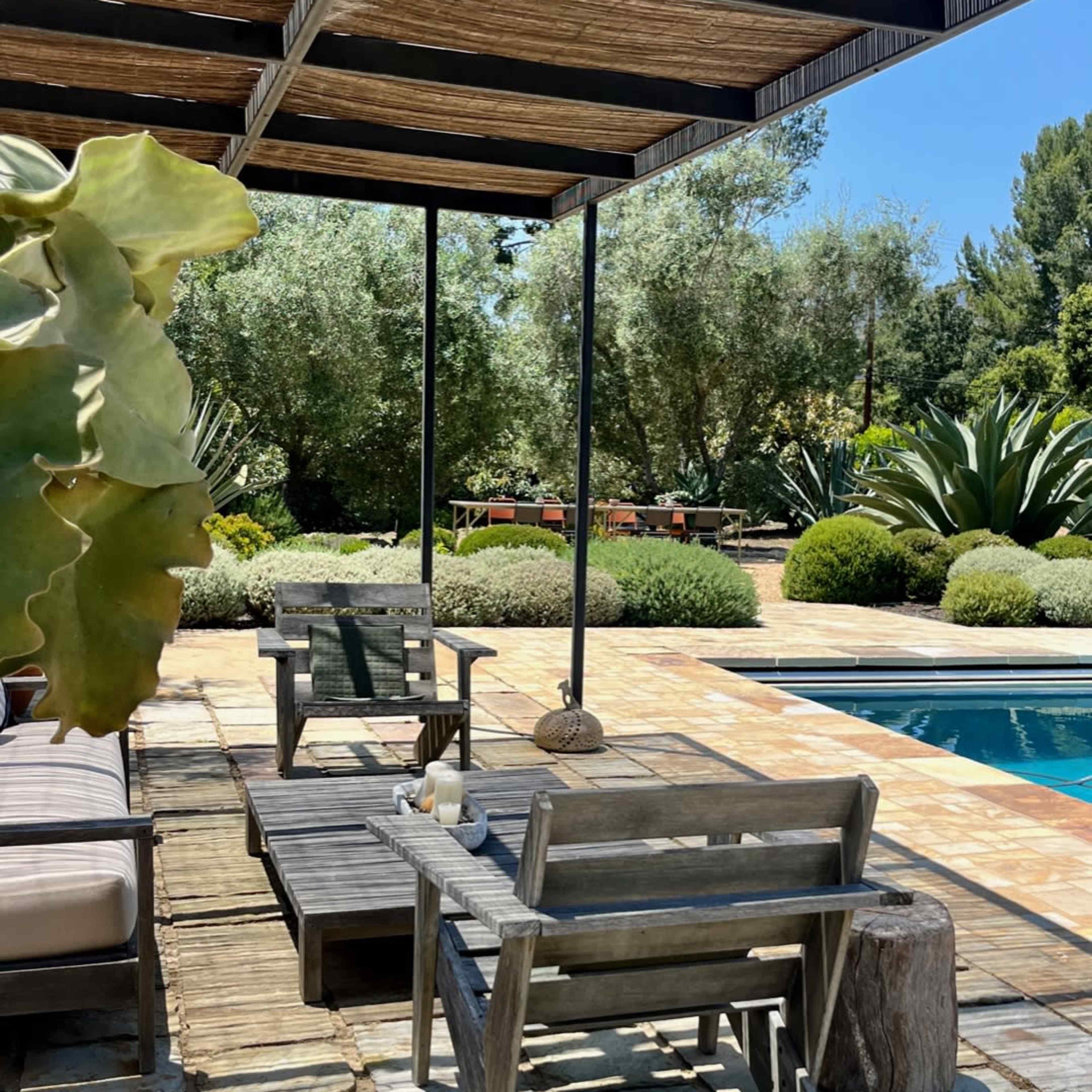A wooden seating area is set beside a pool, surrounded by greenery and shaded by a canopy.