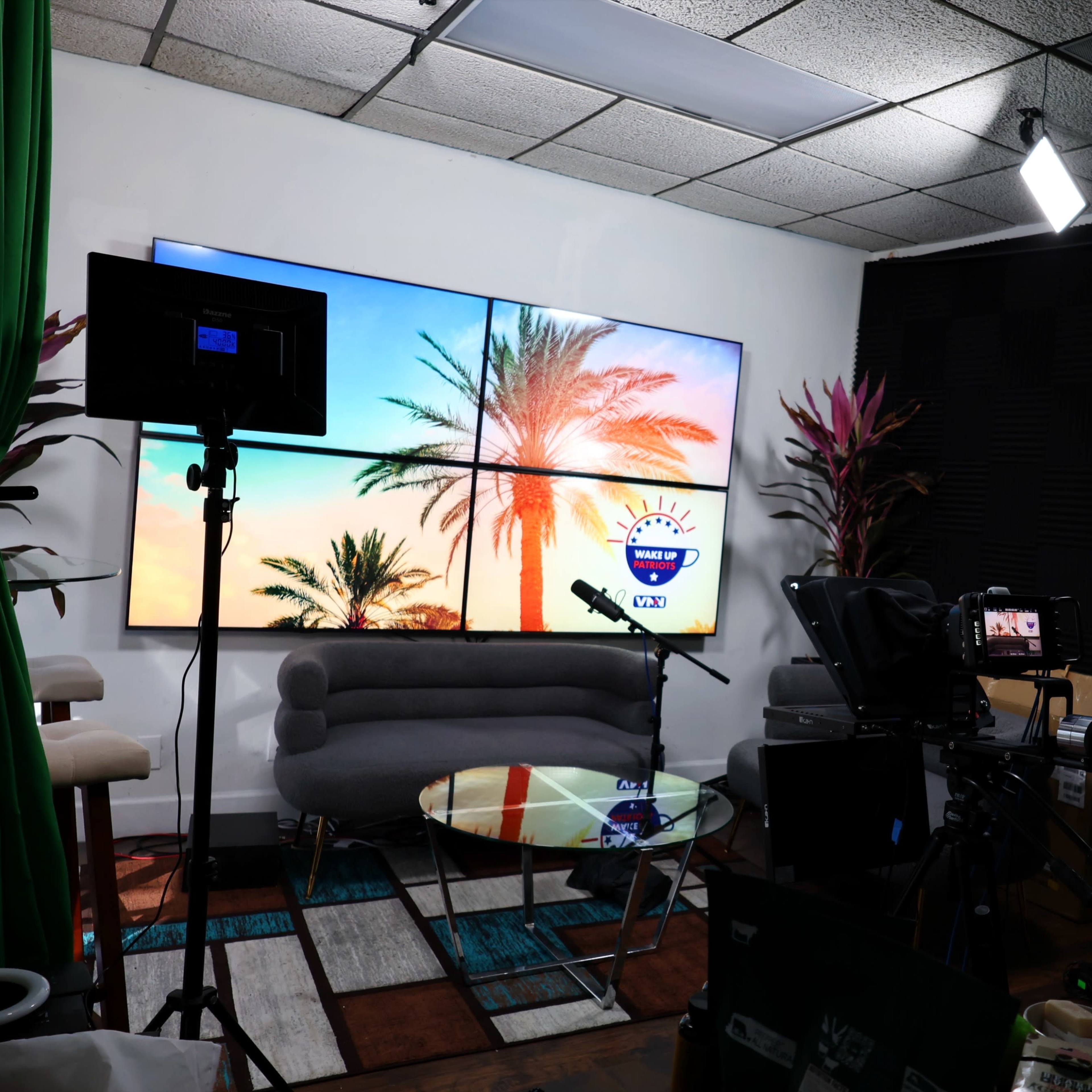 A television studio setup featuring a large screen with palm trees, a round glass table, and multiple cameras and lighting equipment.