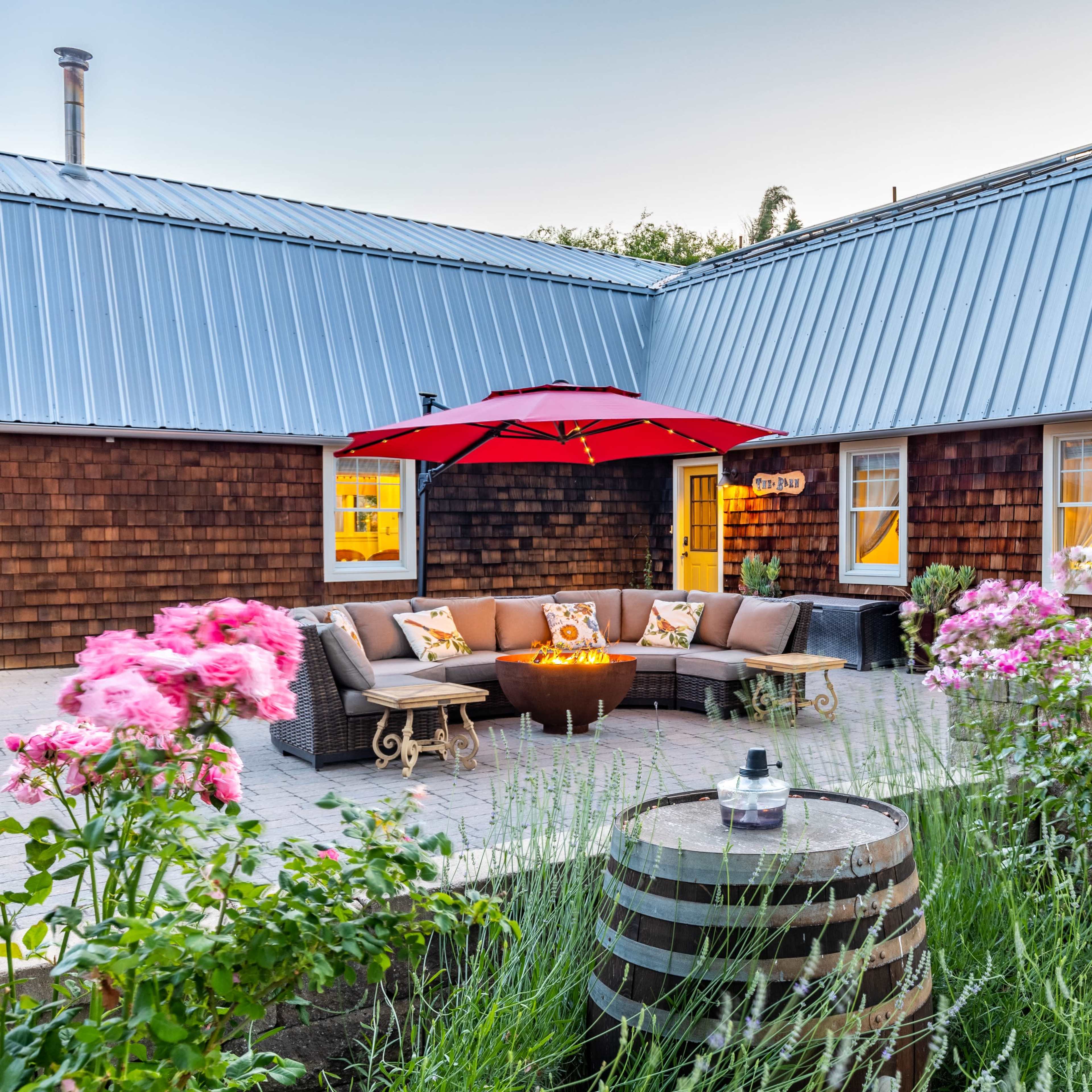 A cozy outdoor seating area with a fire pit is surrounded by flowers and rustic wooden barrels in a courtyard.