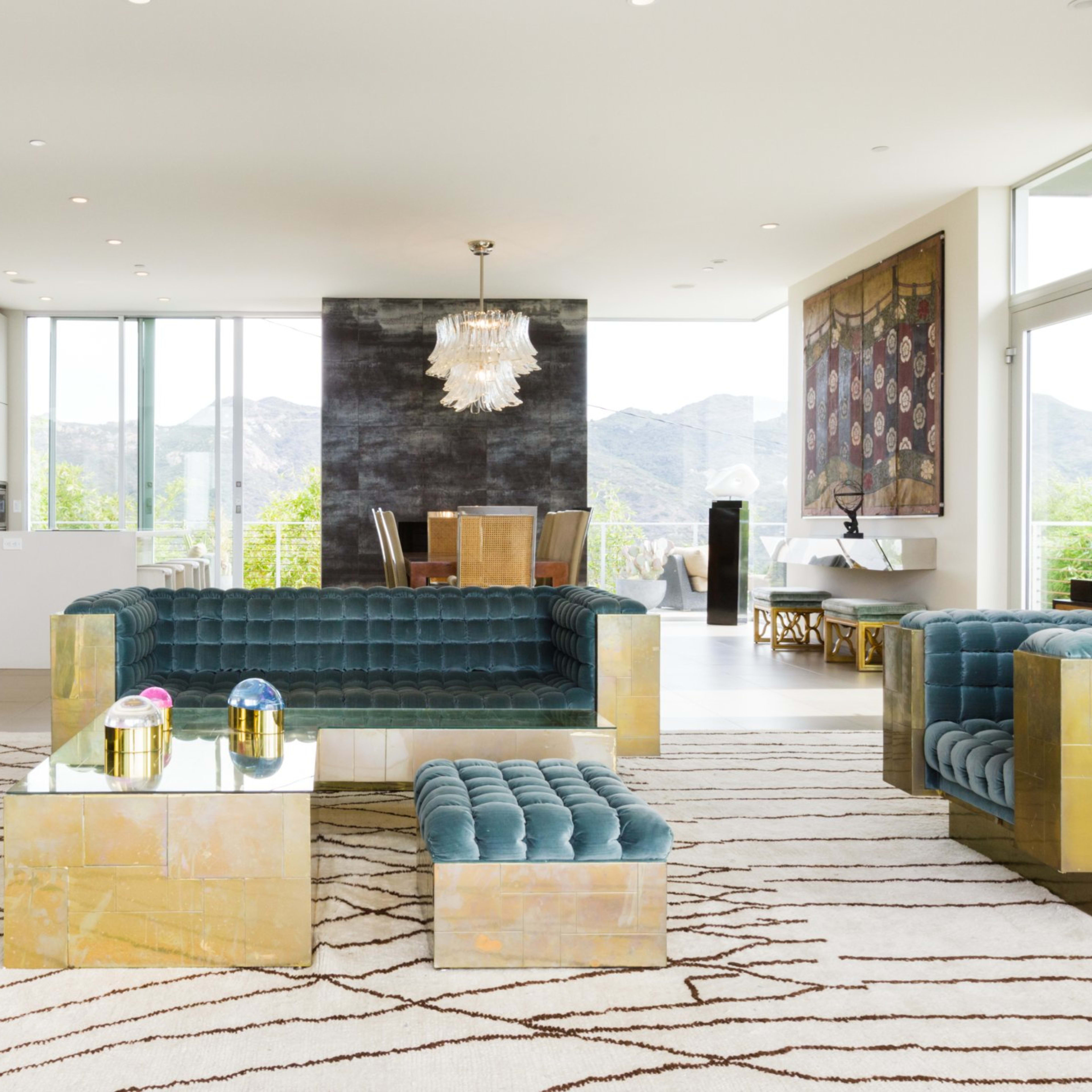 A contemporary living room with teal upholstered couches and gold-accented furniture, overlooking a scenic view through large windows.