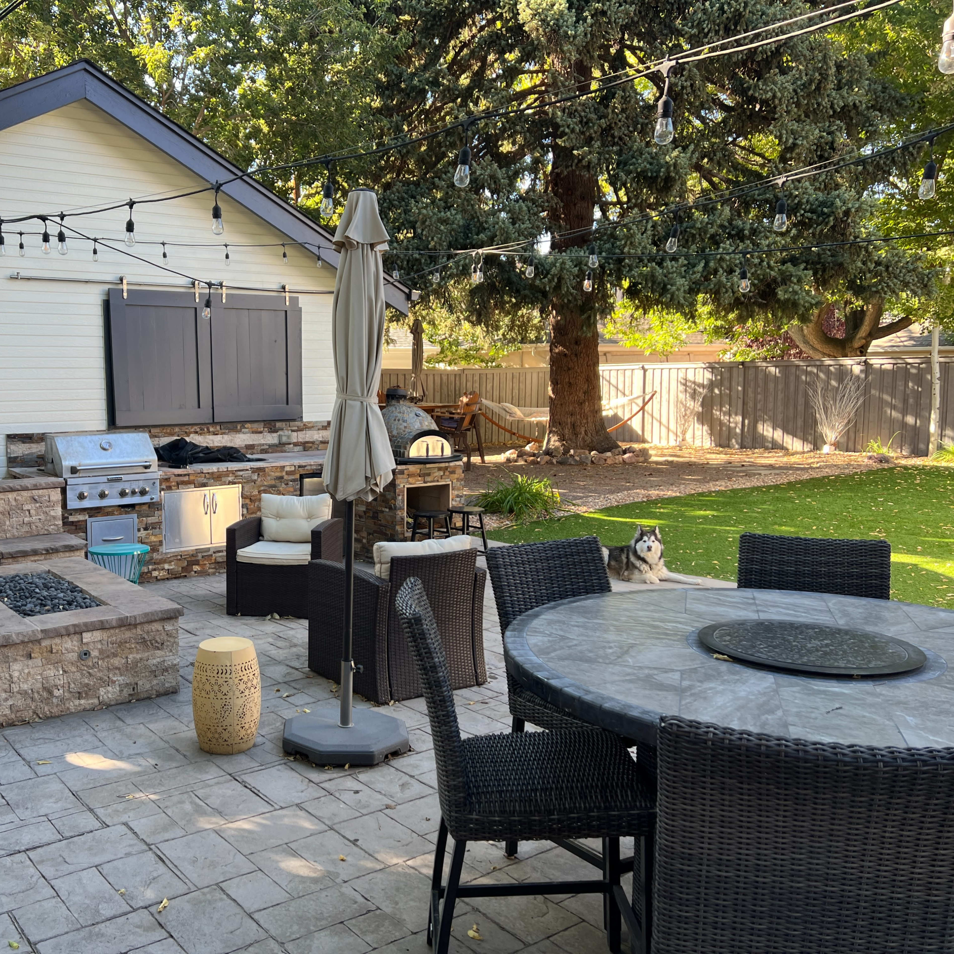 A patio area features a dining table, seating arrangements, a grill, a fire pit, and decorative string lights against a backdrop of a house and trees.