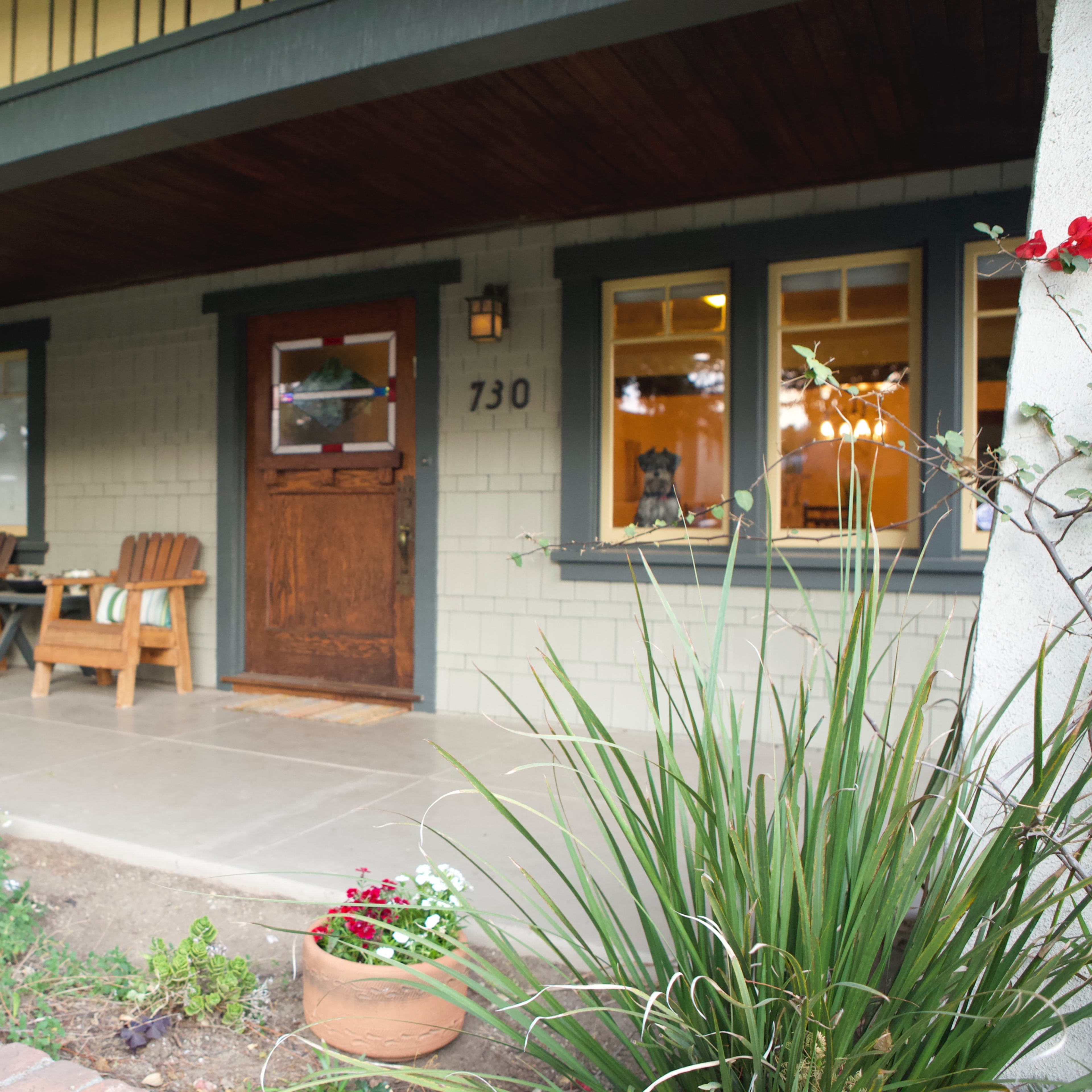 A porch with wooden chairs and a front door is surrounded by potted plants and greenery.