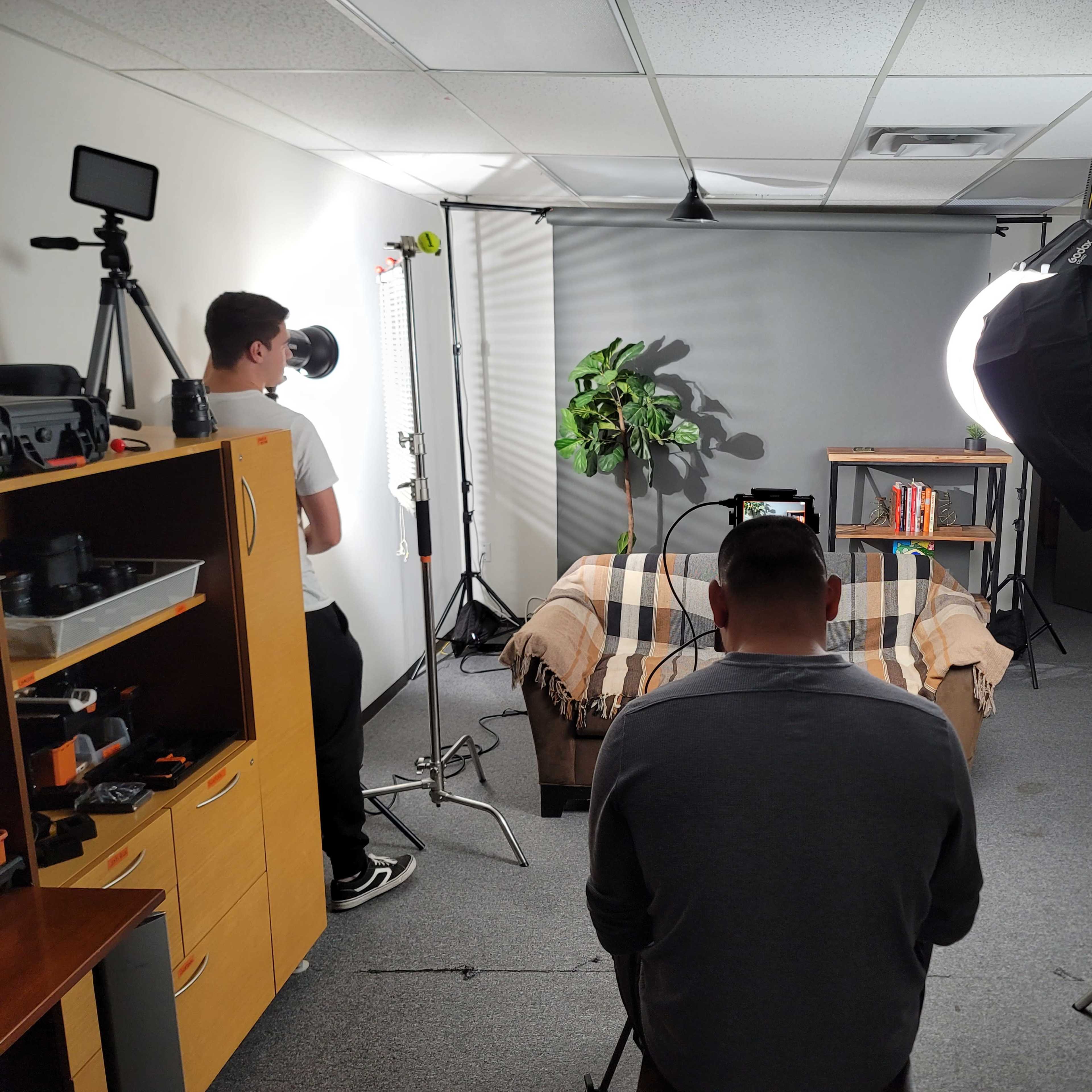A person sits on a couch in a well-lit studio setting with equipment and a backdrop while another person operates a camera.