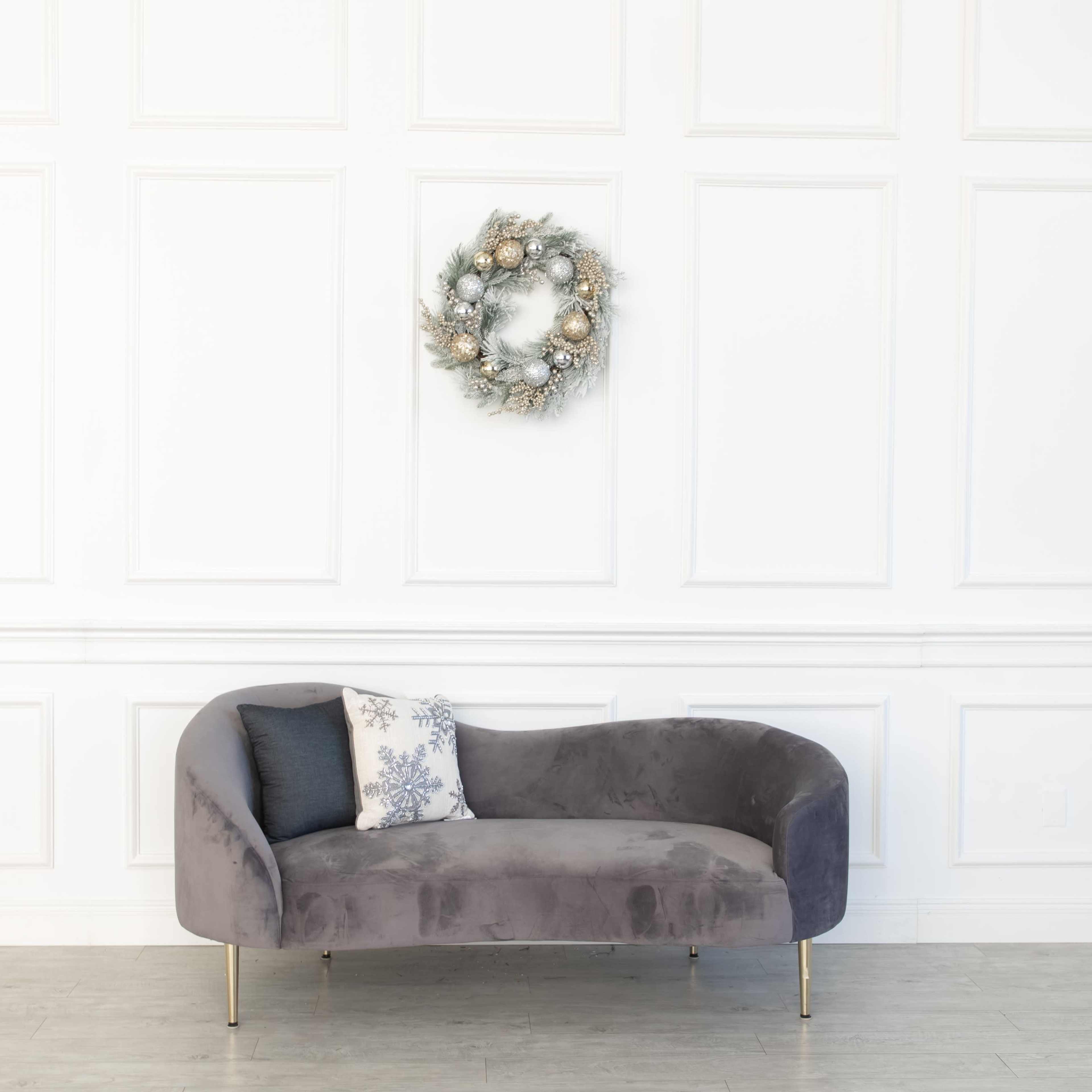 A gray sofa with gold legs is positioned against a white paneled wall, adorned with a decorative wreath above it.
