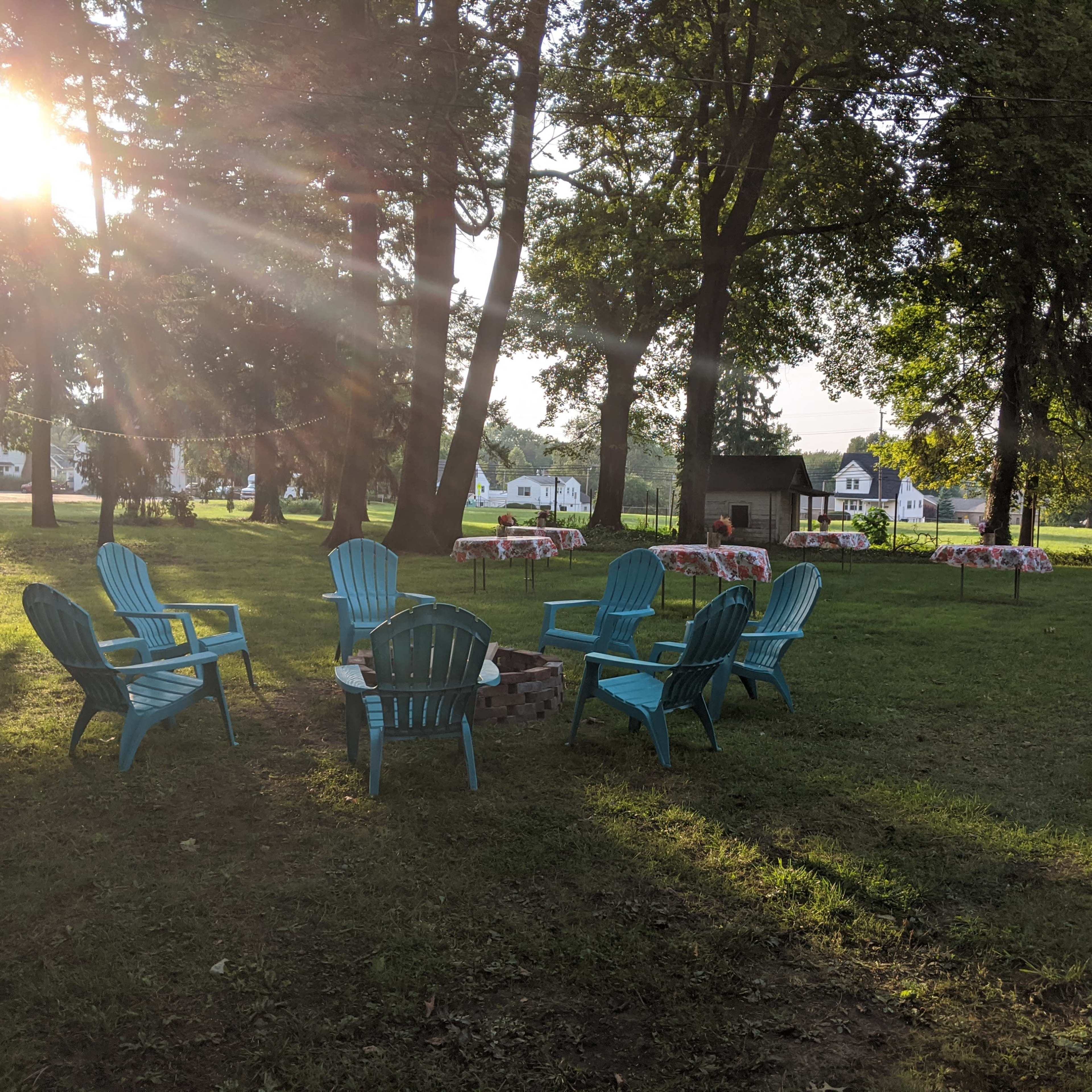 A cluster of blue Adirondack chairs surrounds a fire pit in a grassy area with tables set up in the background.