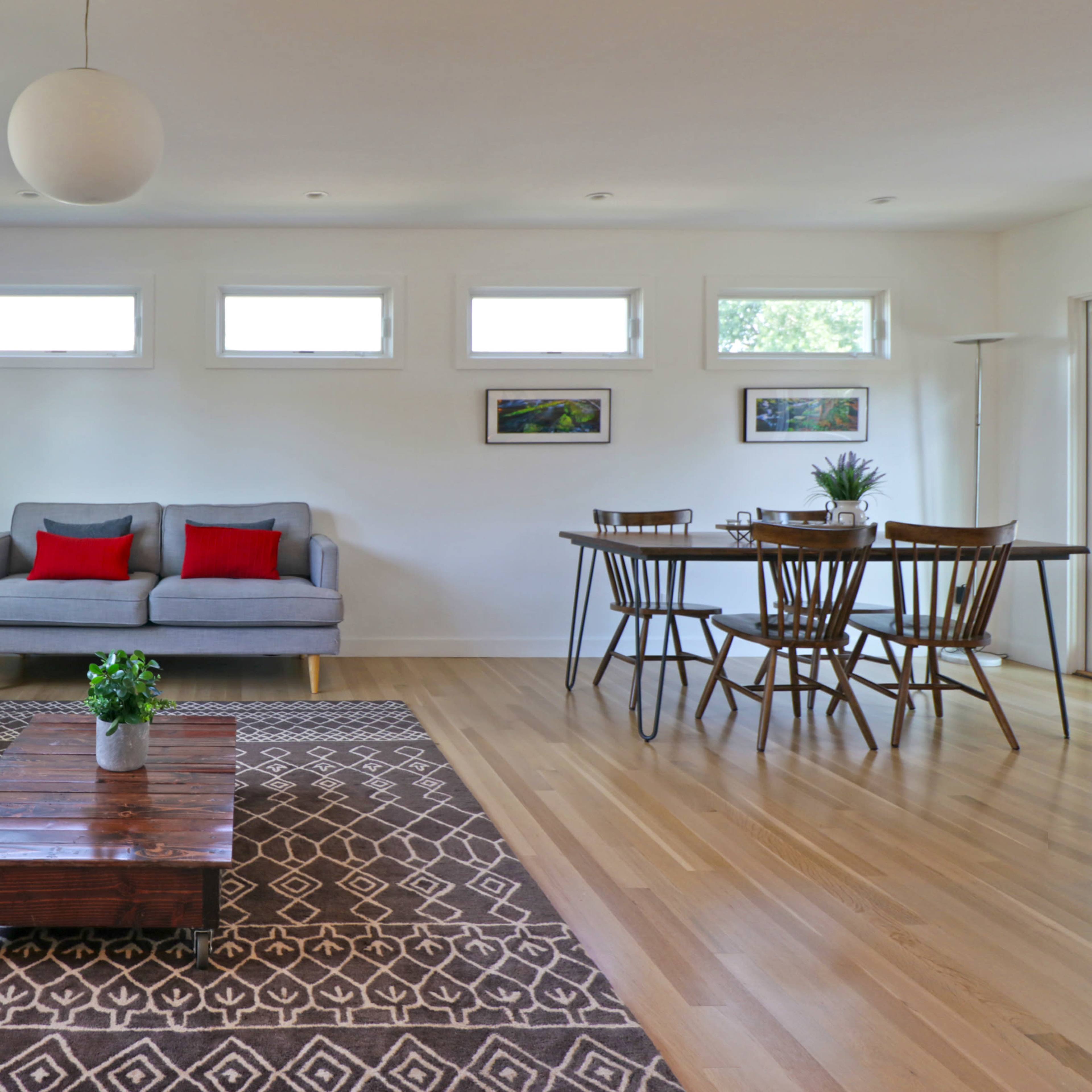A modern living room features a gray sofa with red cushions, a wooden coffee table, a dining area with a table and chairs, and large windows that allow natural light.