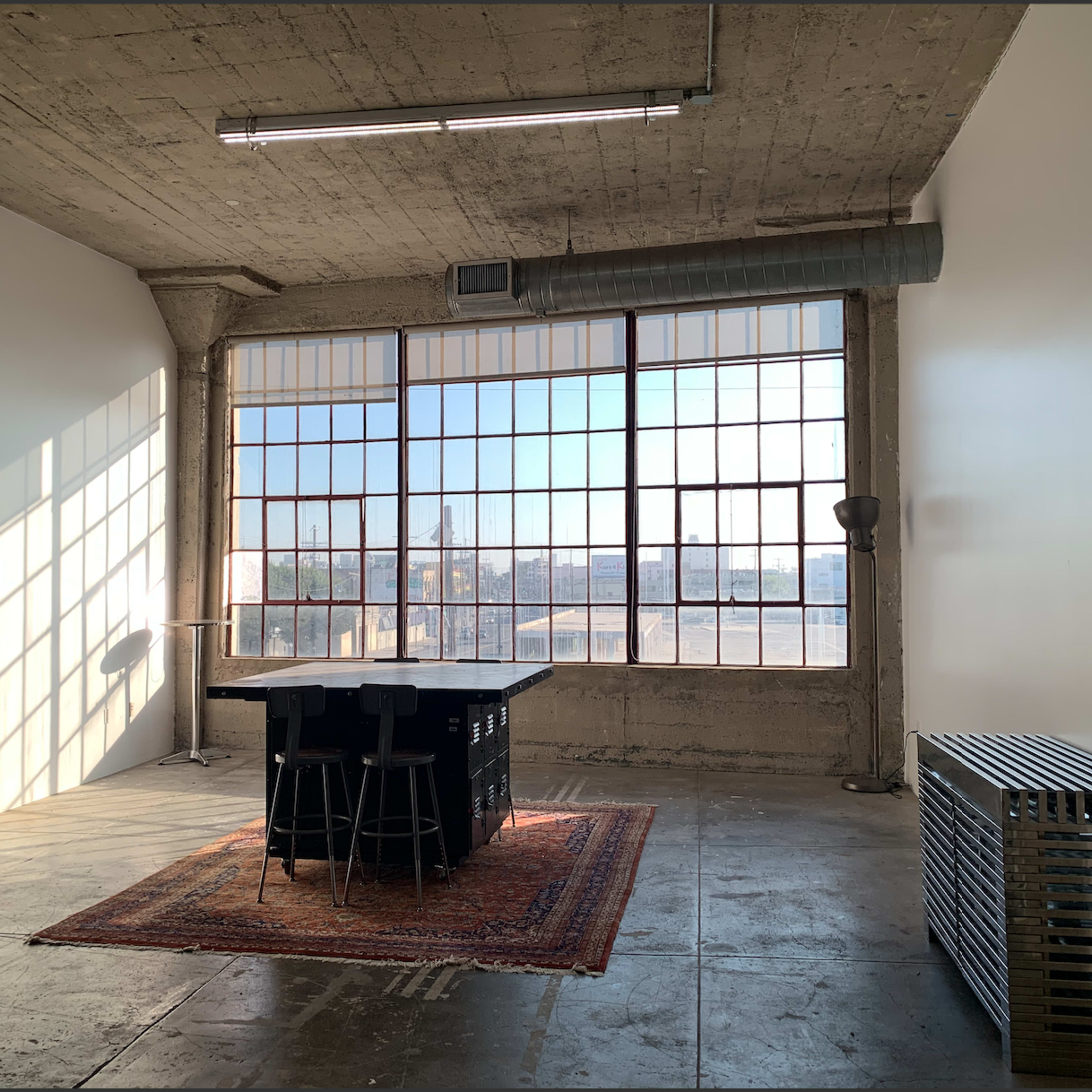 A spacious room with large windows, concrete walls, a rug, a table with stools, and an arcade machine in the corner.