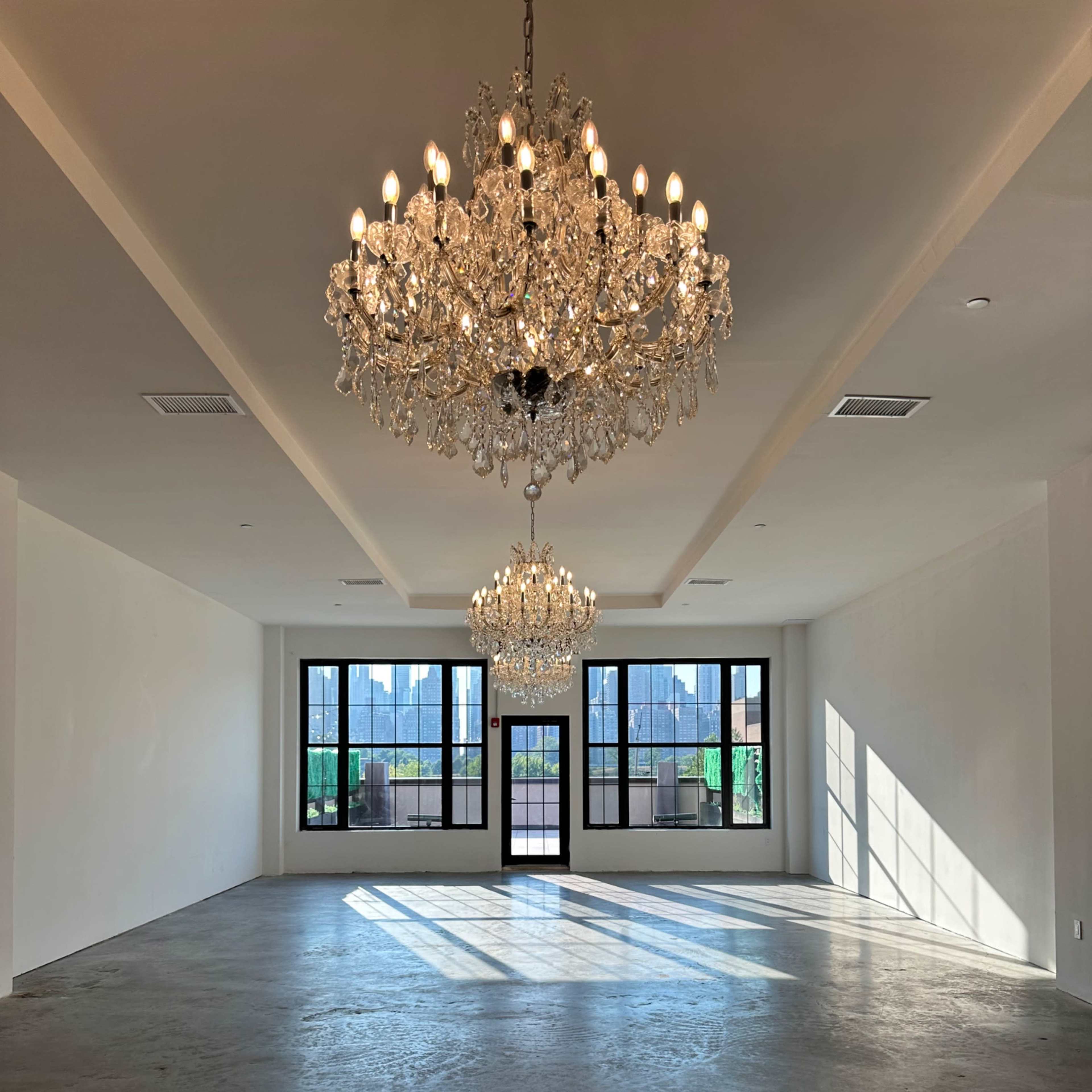 A spacious room featuring two large chandeliers and several large windows that cast shadows on the concrete floor.