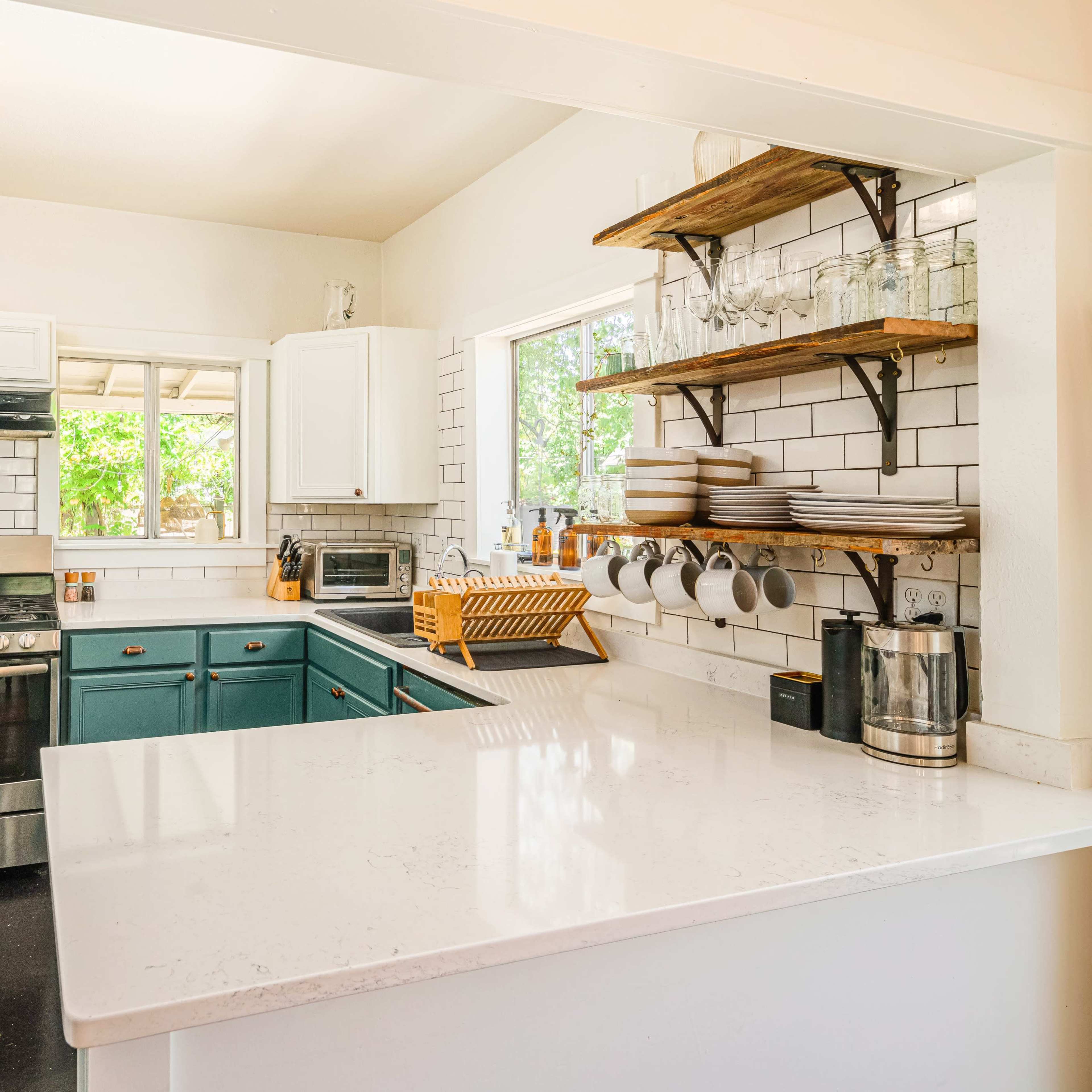 A modern kitchen with teal cabinets, white countertops, and open shelving displaying dishes and glassware.
