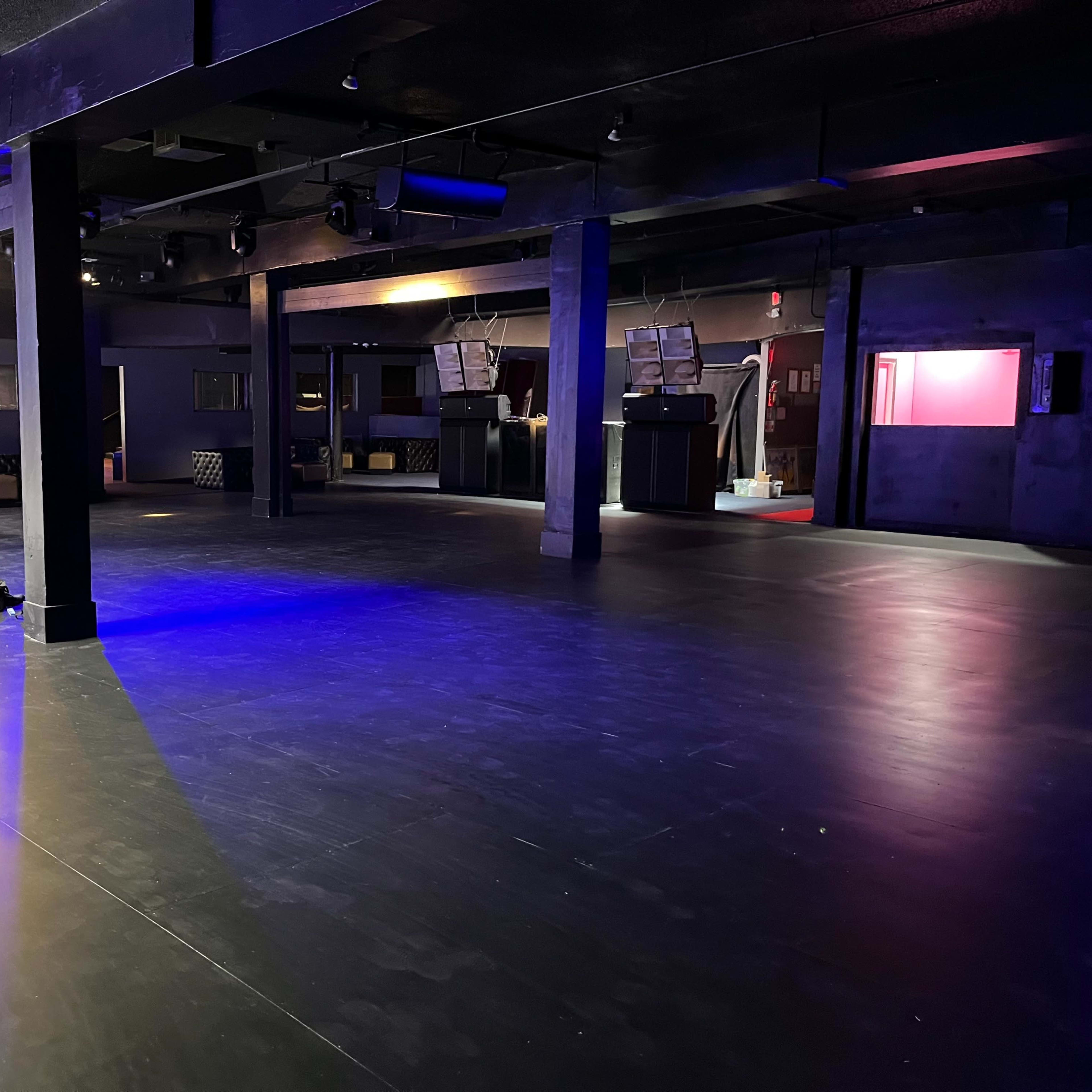 A spacious, dimly lit interior of a nightclub with black flooring and some construction tools scattered on the ground.