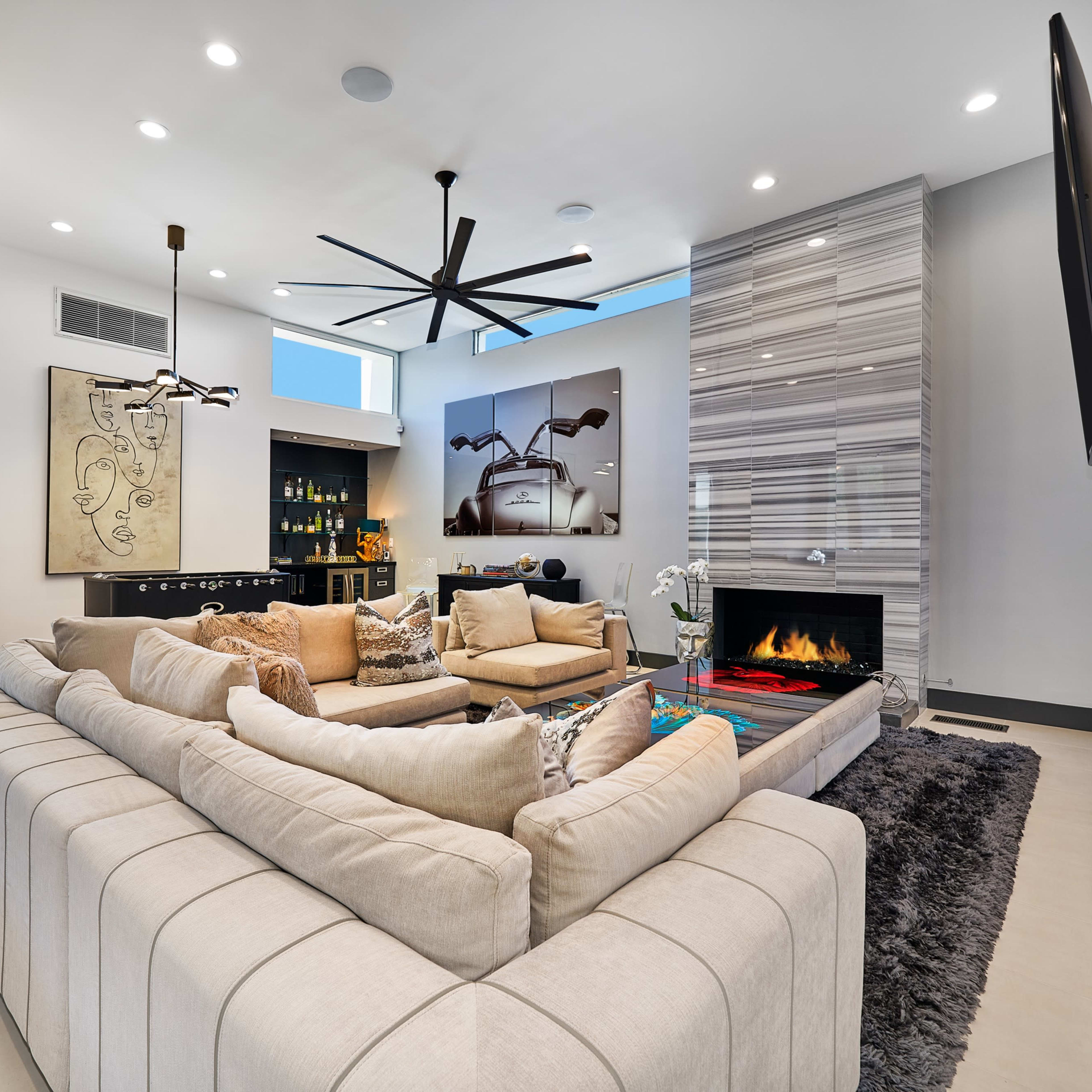 A modern living room with a sectional sofa, a wall-mounted television, a decorative fireplace, and large windows allowing natural light to enter.
