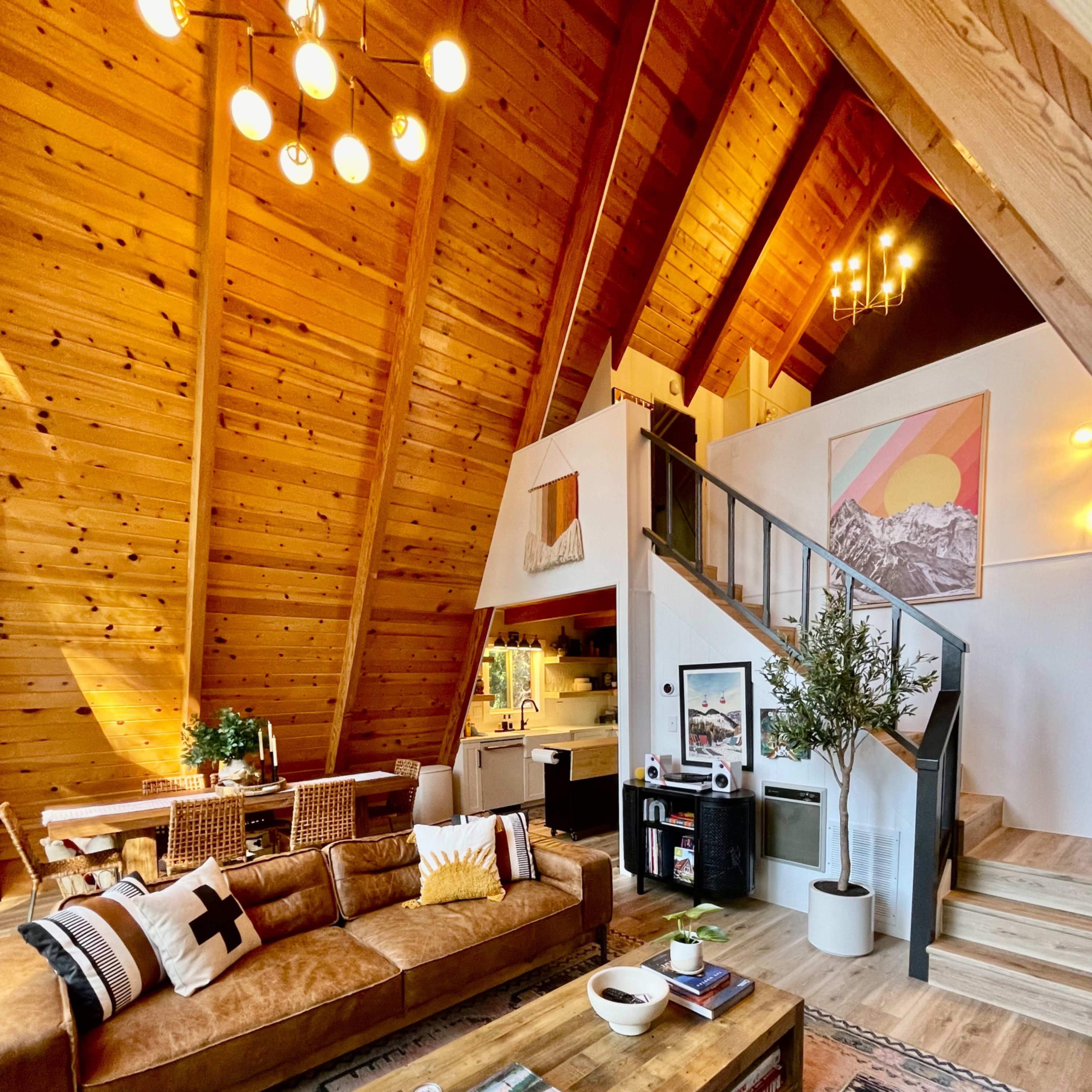 A cozy, wooden A-frame living room featuring a brown leather sofa, a large coffee table, and a staircase leading to the upper level.