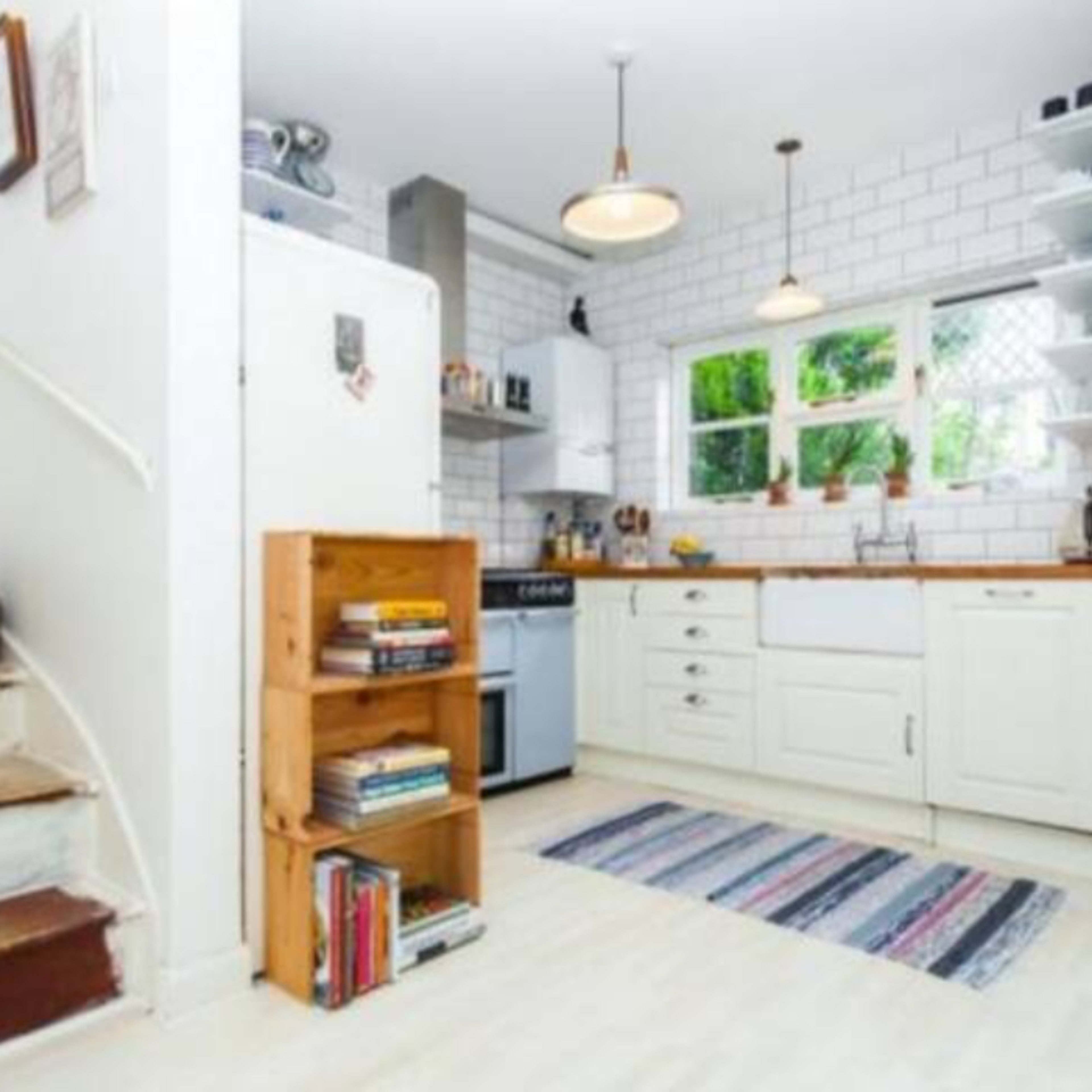 A bright kitchen with white tile walls, wooden cabinets, a large window, and a small bookshelf filled with books beside a staircase.