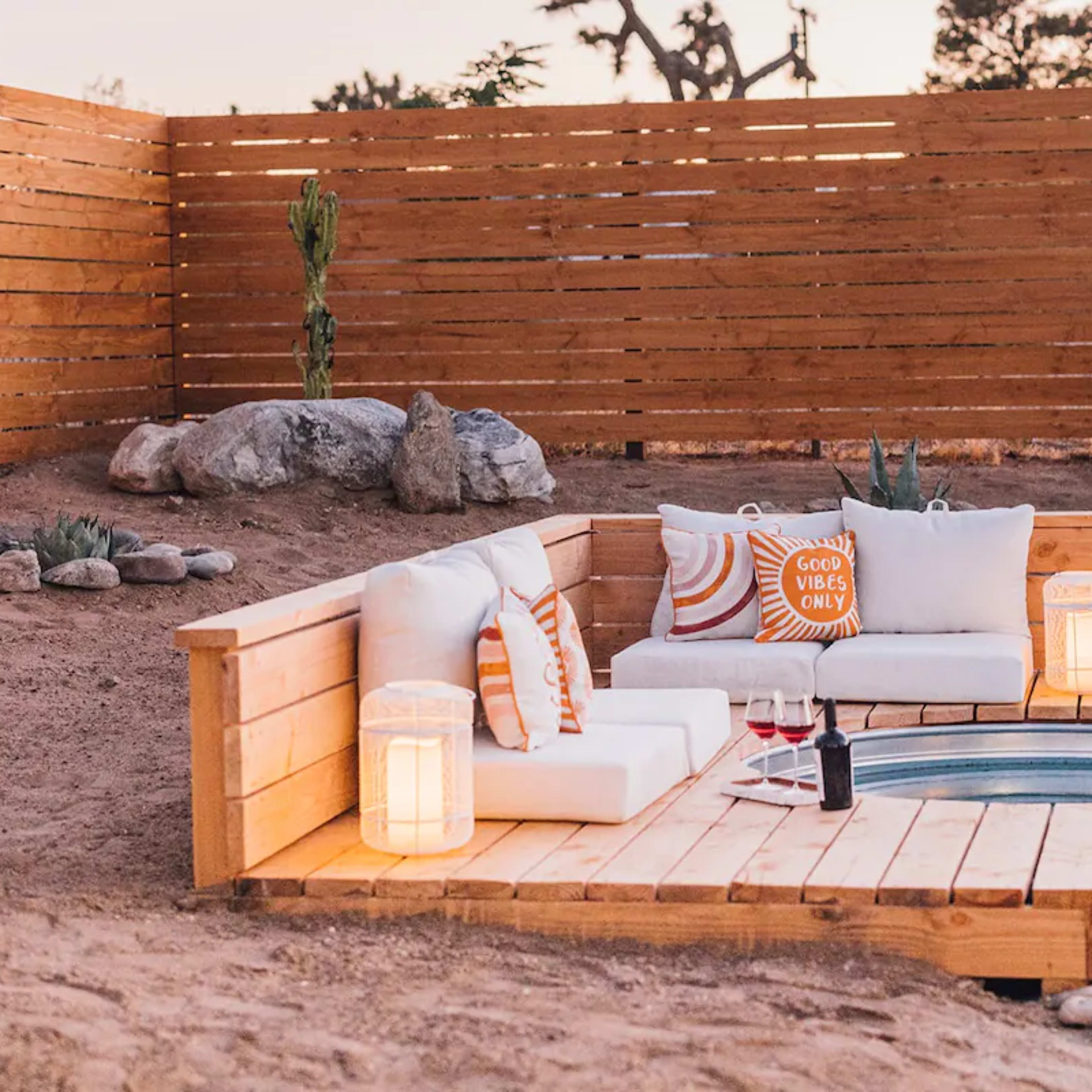 A wooden seating area is set up beside a circular hot tub, featuring white cushions and two lanterns, in a desert landscape.
