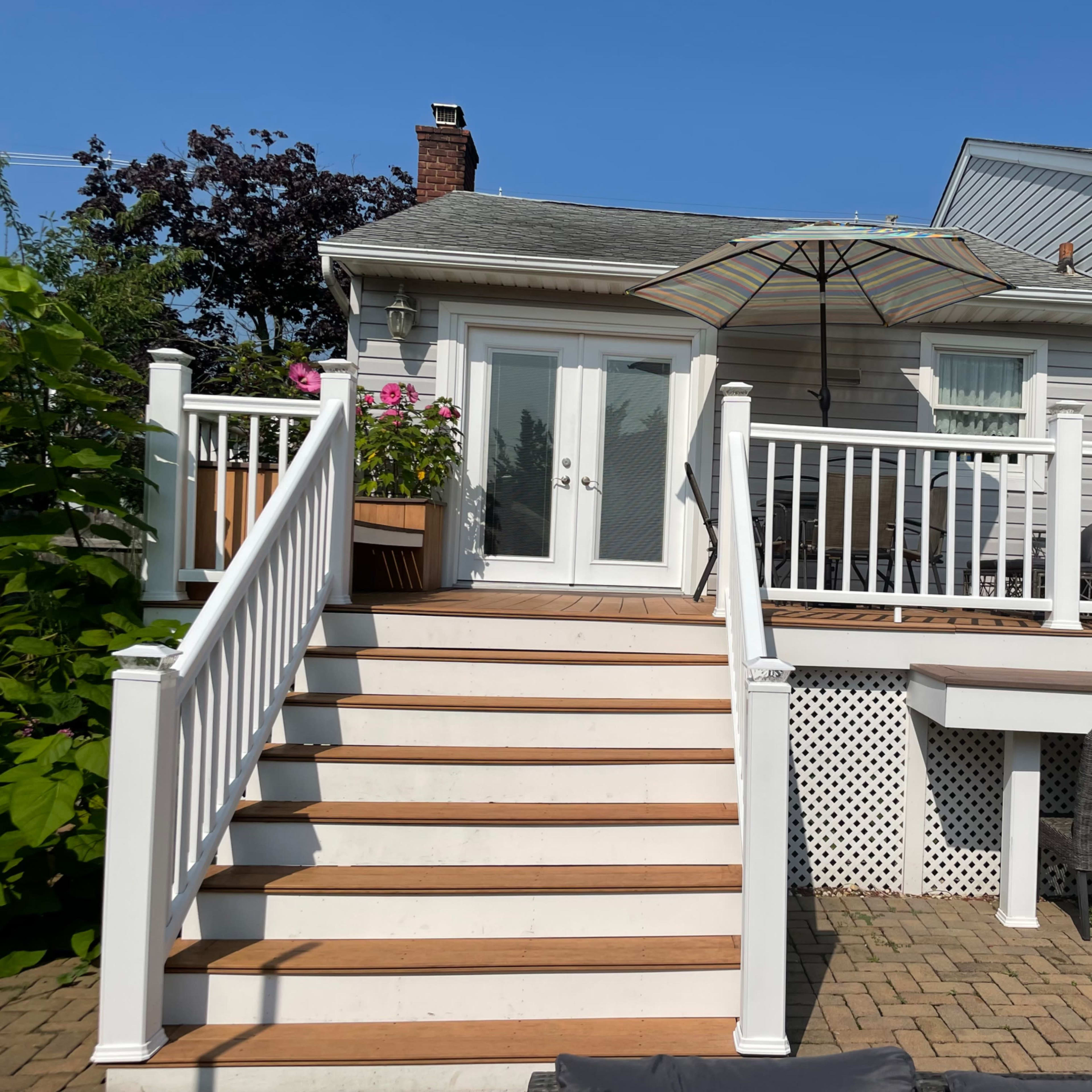 A set of wooden stairs leads up to a deck with white railings and an umbrella, adjacent to a house with a stone and siding exterior.