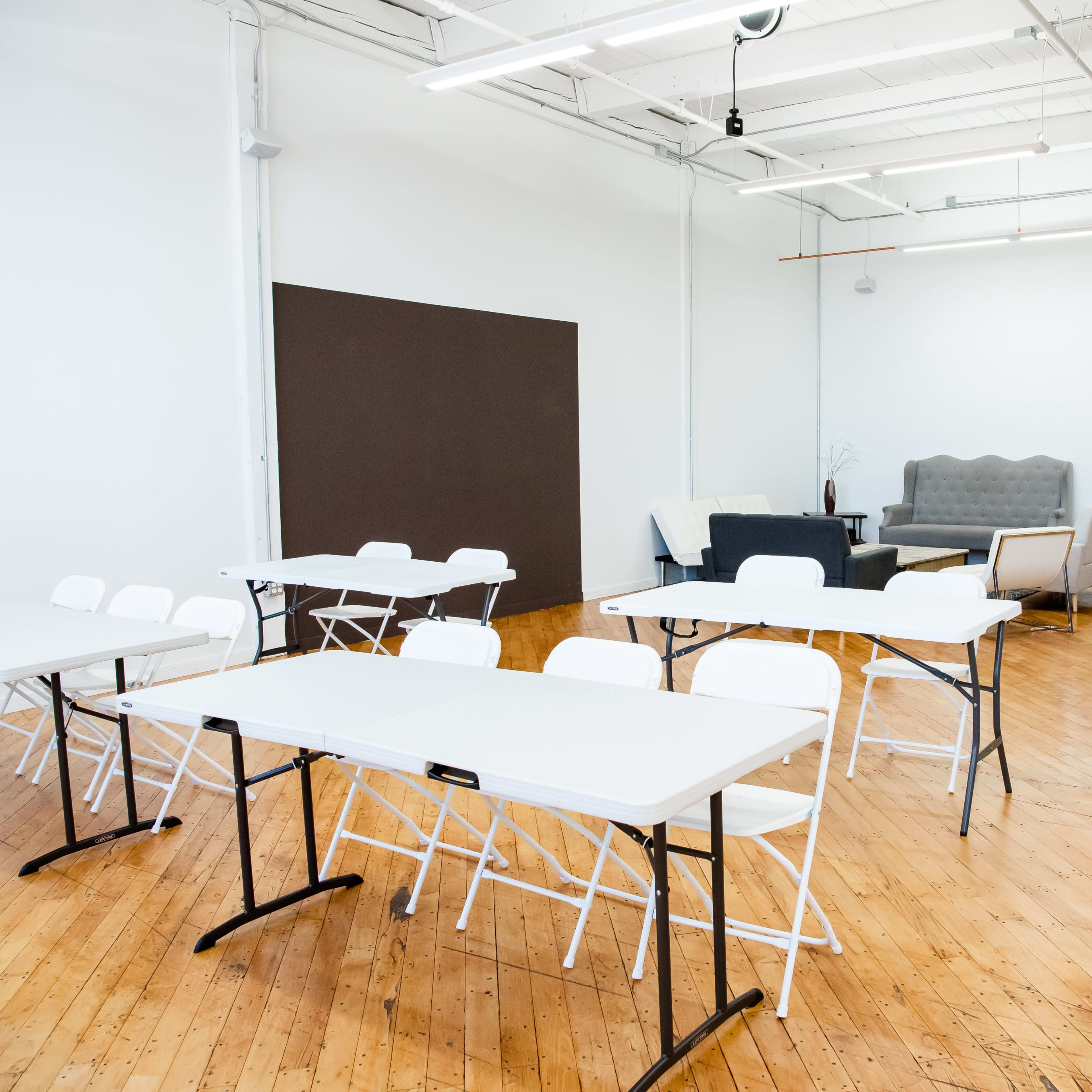 A spacious, well-lit room with several white folding tables and chairs arranged in a simple layout, alongside a seating area with a couch and a kitchenette in the background.