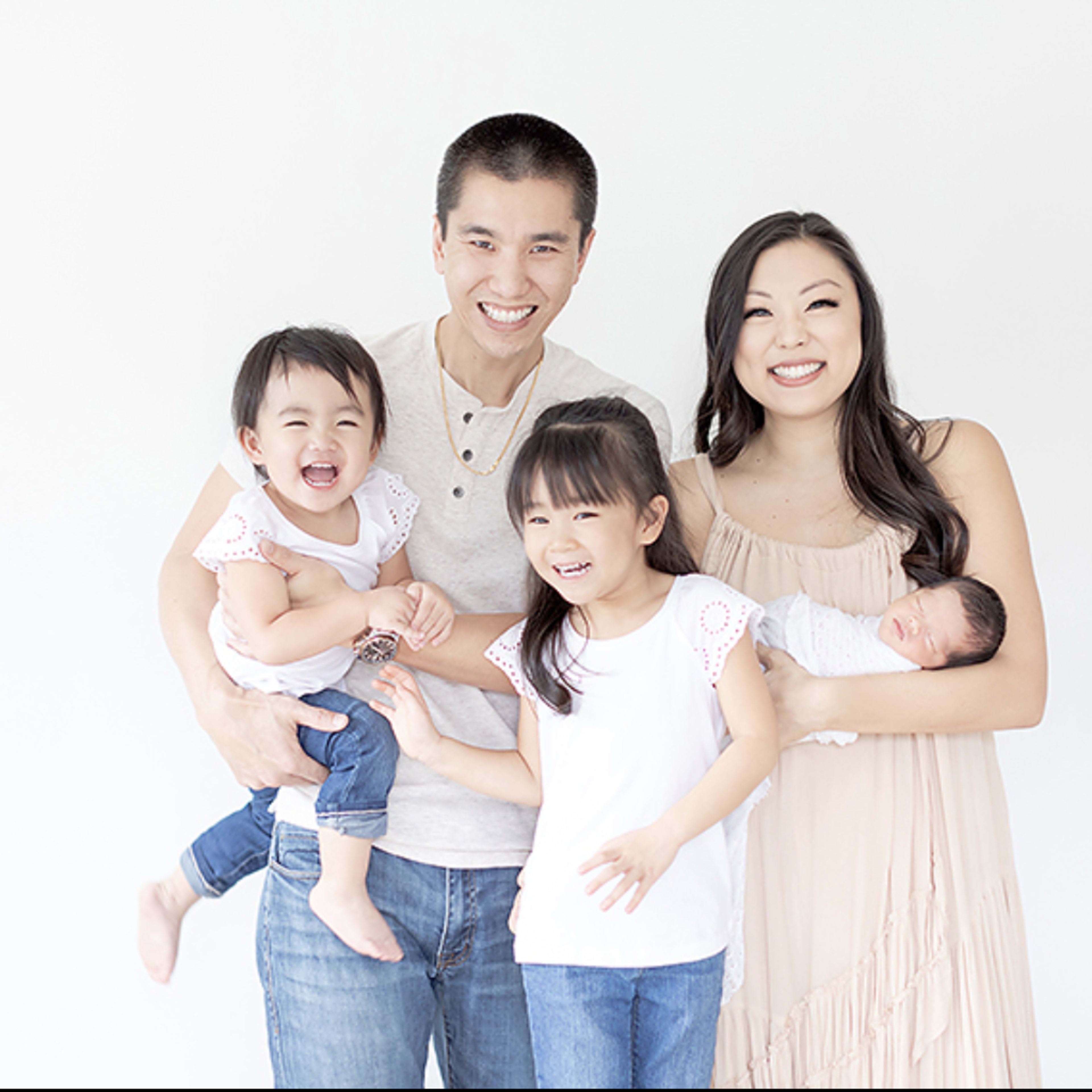 A family of five stands together, with two parents holding their three young children against a plain white background.