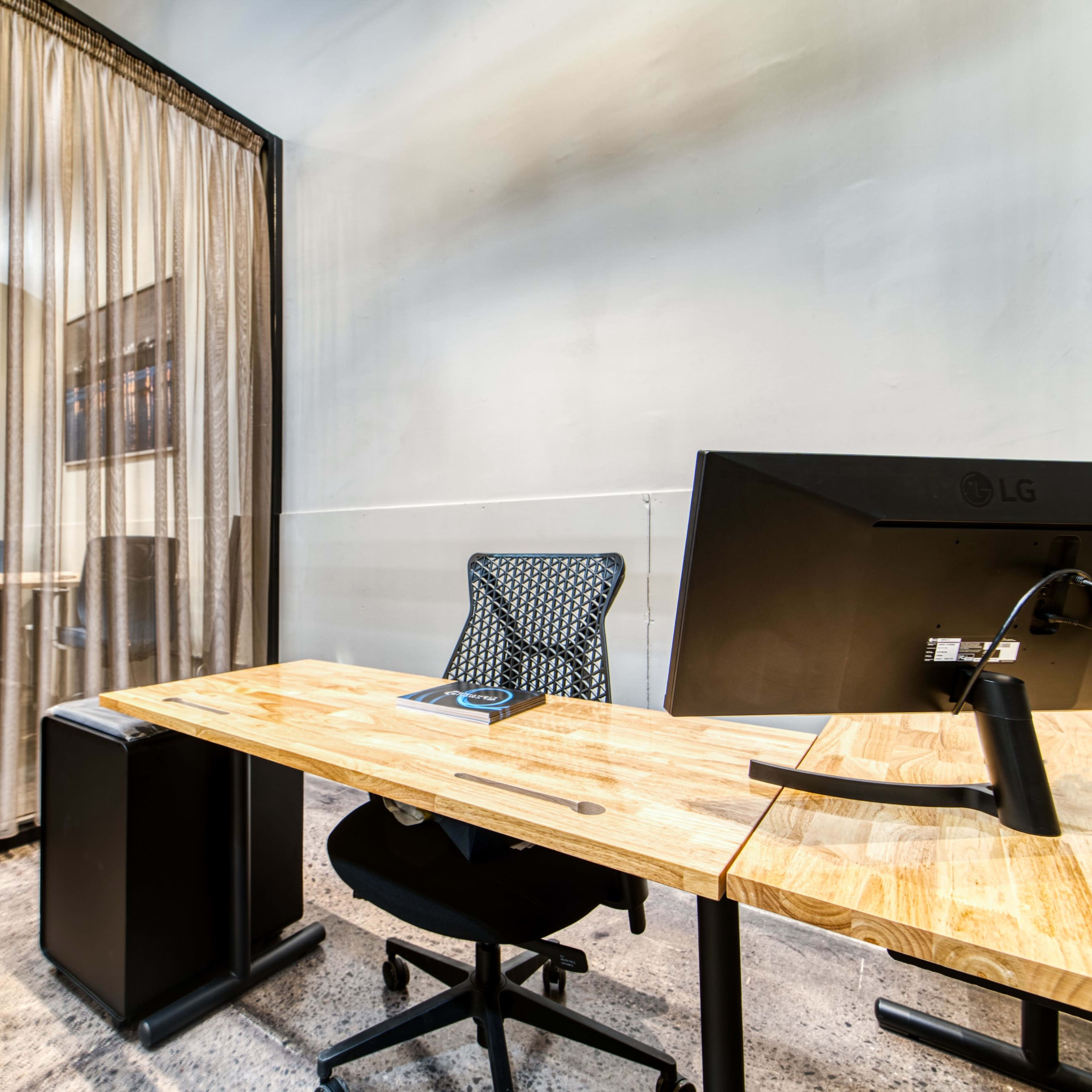 A modern office workspace featuring a wooden desk, an ergonomic chair, and a large computer monitor, separated by a sheer curtain from a second workspace area.