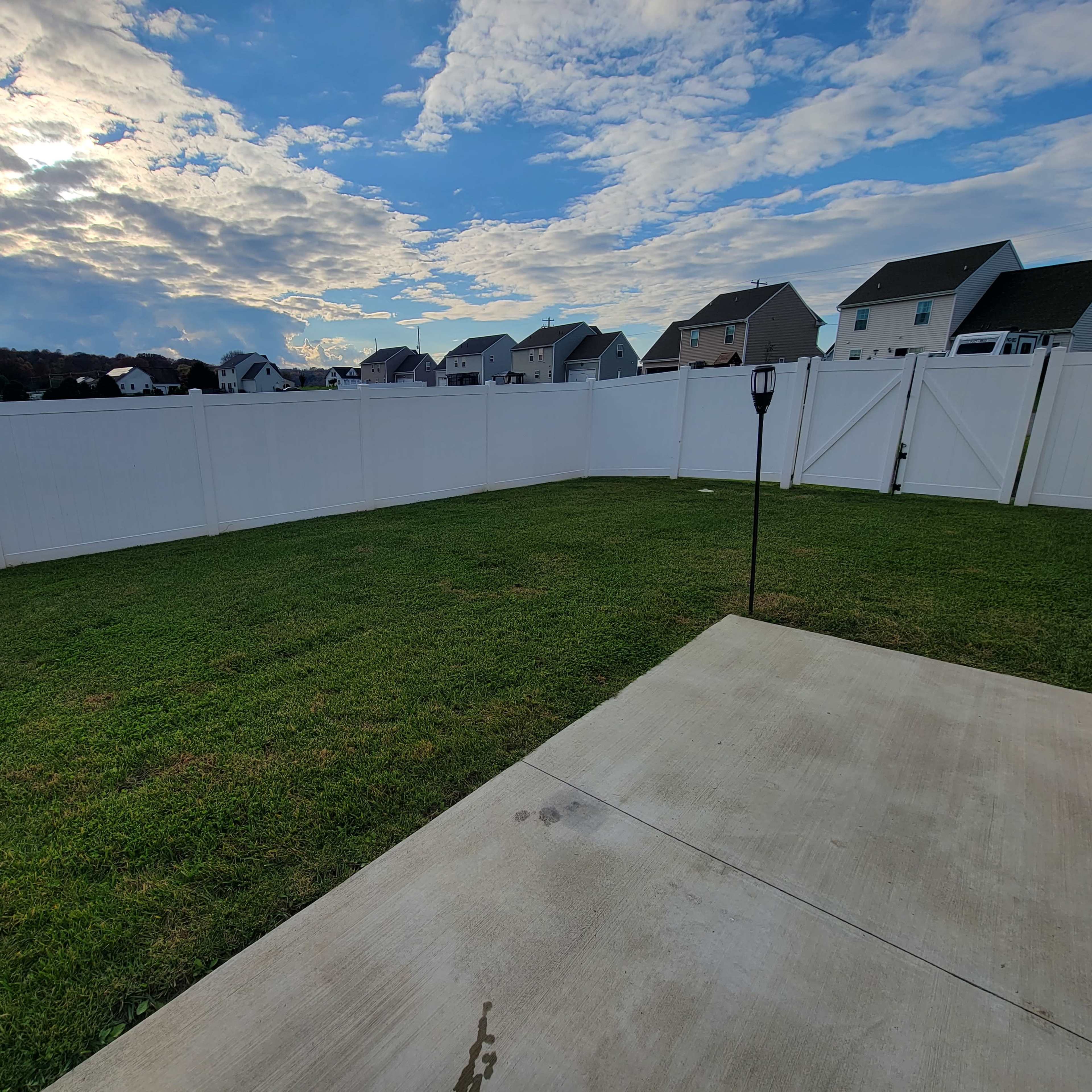 A grassy backyard enclosed by a tall white fence, with a concrete patio area and houses visible in the background under a partly cloudy sky.
