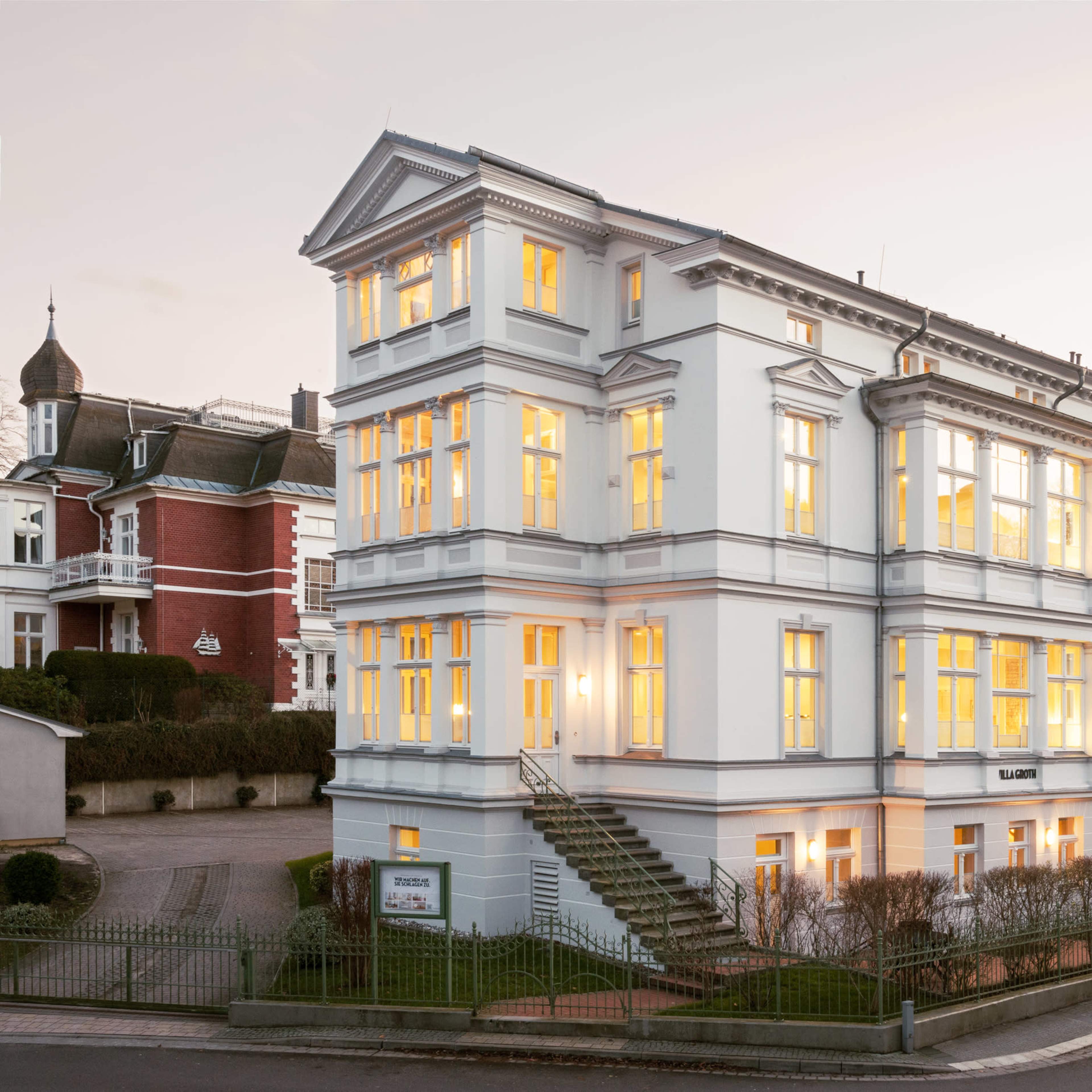 A three-story white building with large windows is illuminated at dusk, situated on a corner surrounded by other structures.