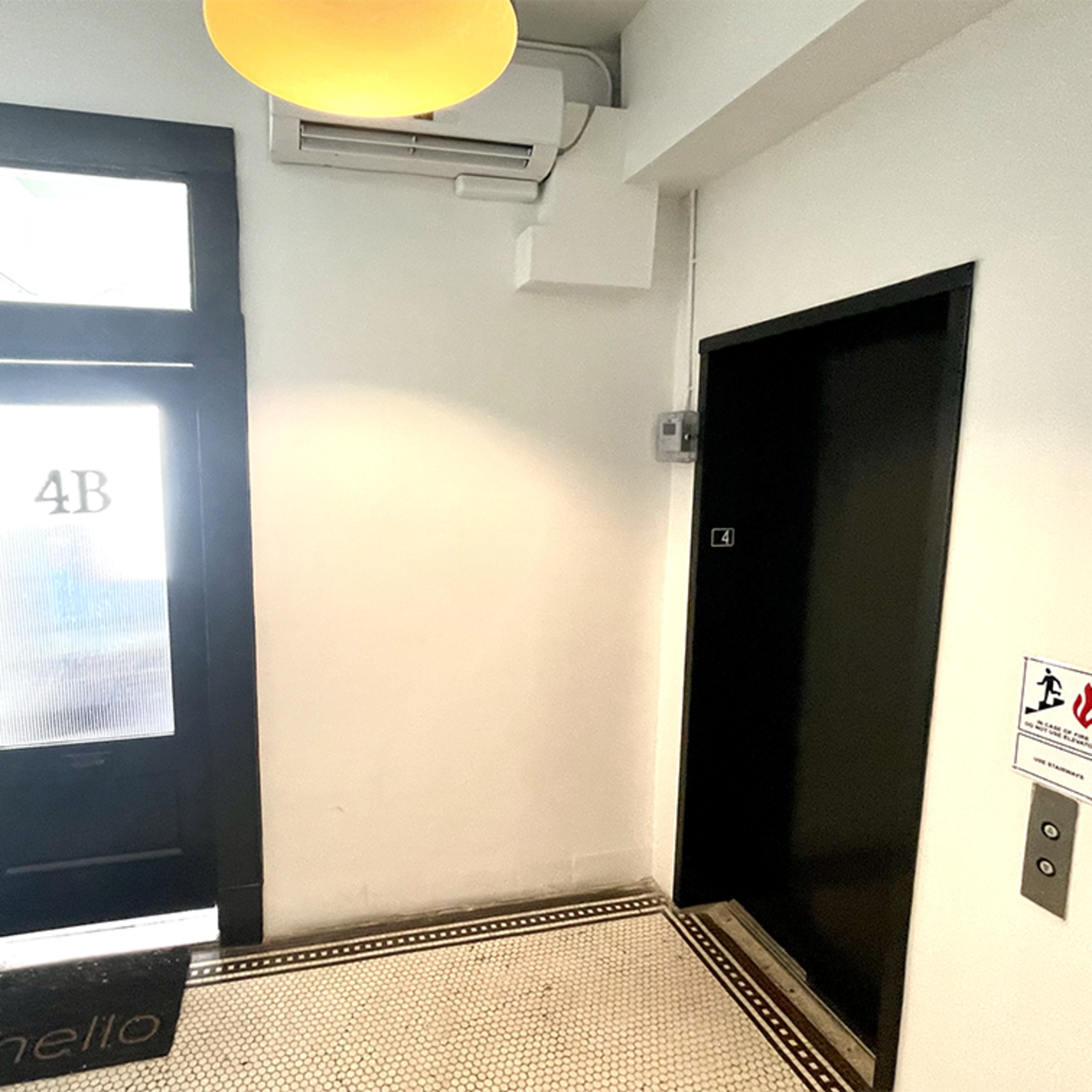 An entryway with a black door labeled "4B," a small welcome mat, an elevator door, and a light fixture on the ceiling.
