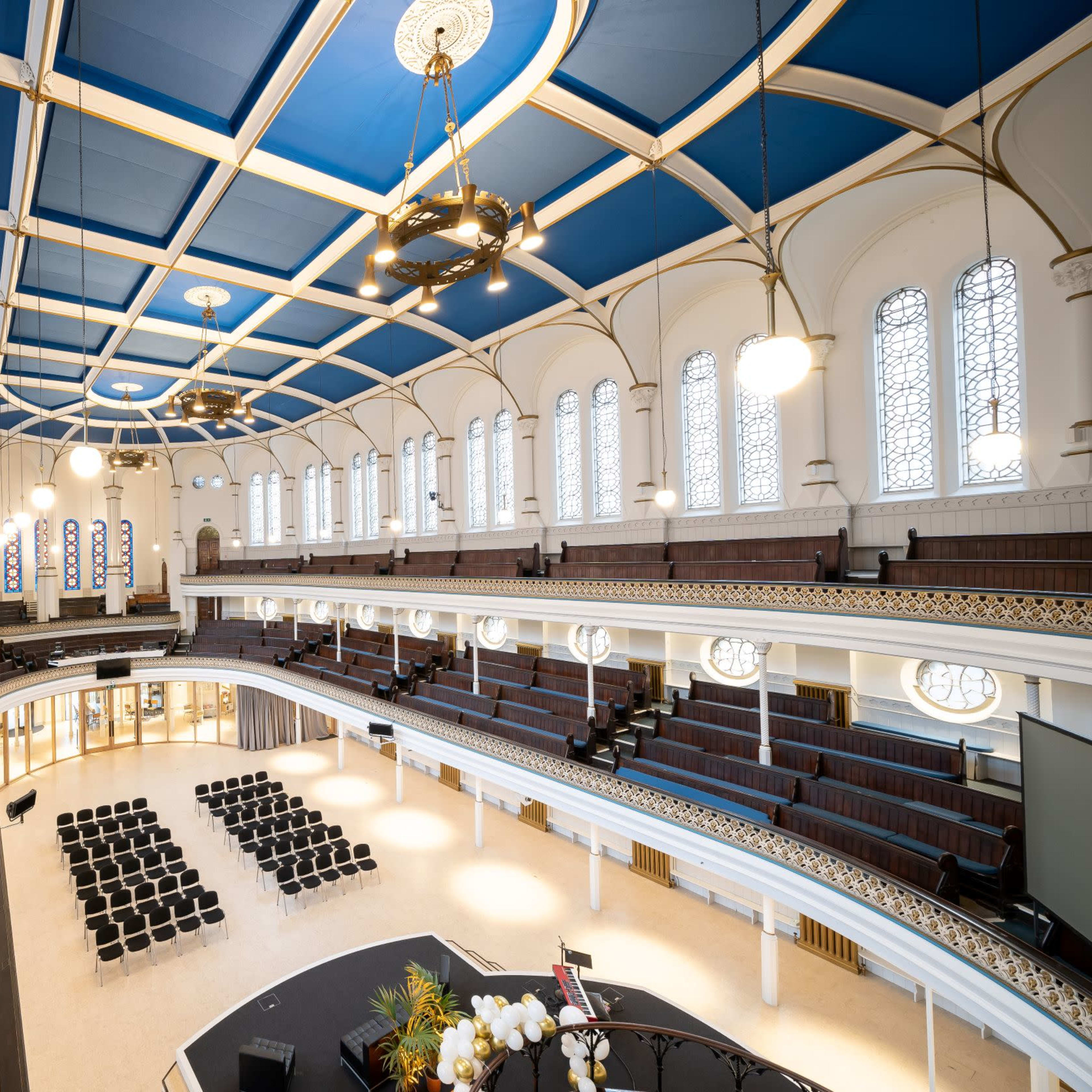A spacious auditorium with a blue ceiling, multiple levels of seating, and large windows letting in natural light.
