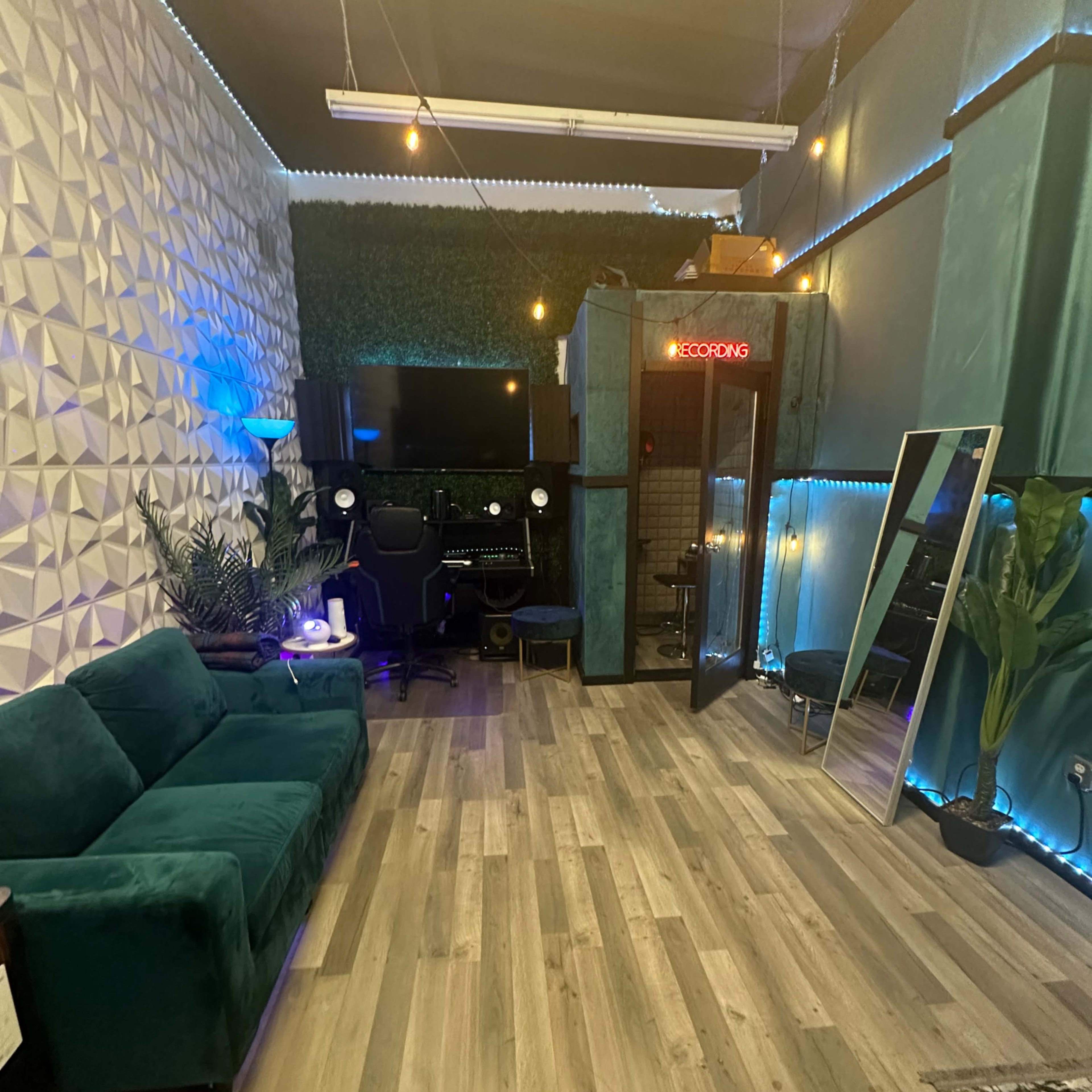 A modern recording studio featuring a teal couch, a workstation with a computer, and a soundproof booth enclosed with glass.