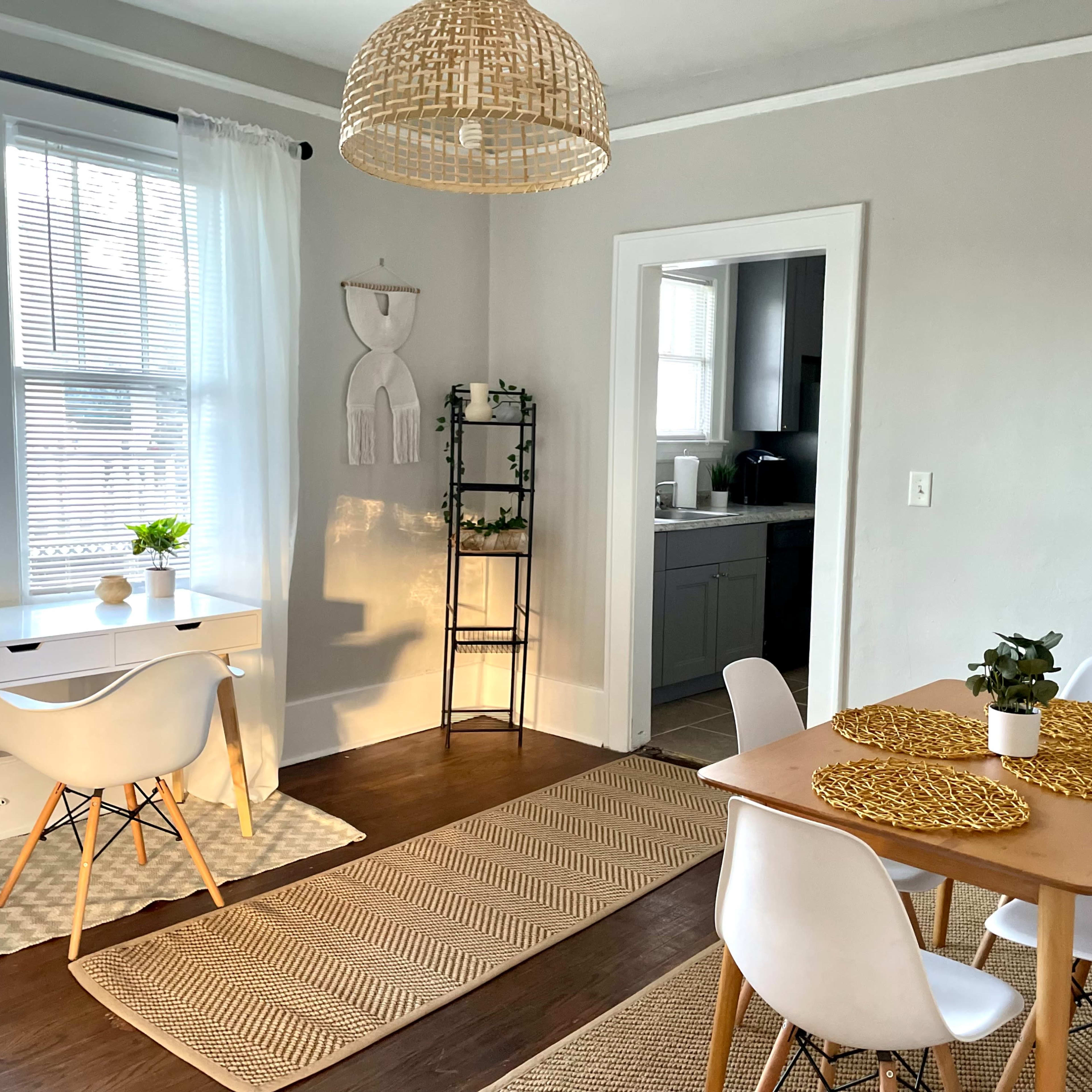 A bright dining area features a wooden table with white chairs, a desk with a plant, and a woven light fixture hanging from the ceiling.