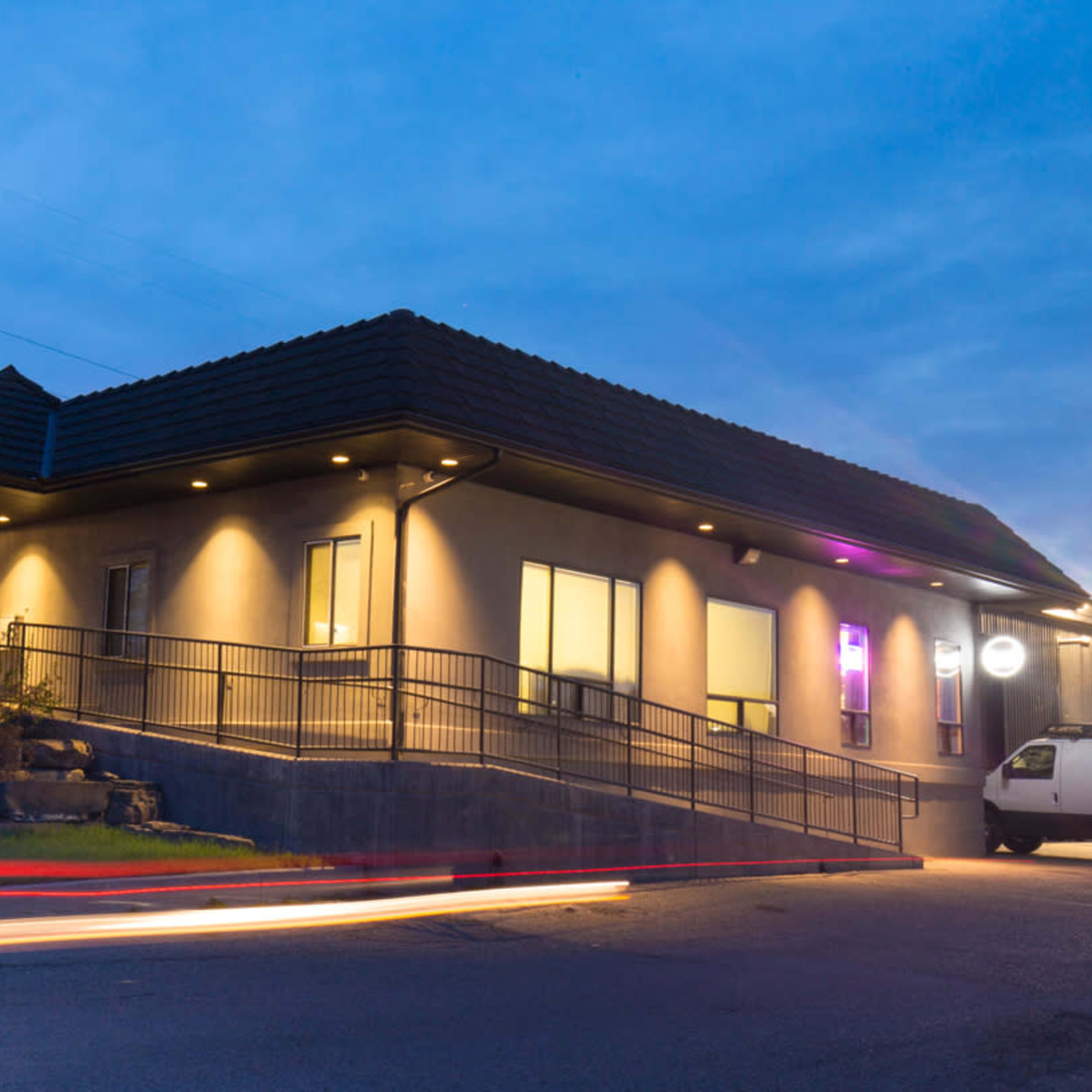 A modern building with a sloped roof, illuminated windows, and a ramp leading to the entrance, set against a twilight sky.