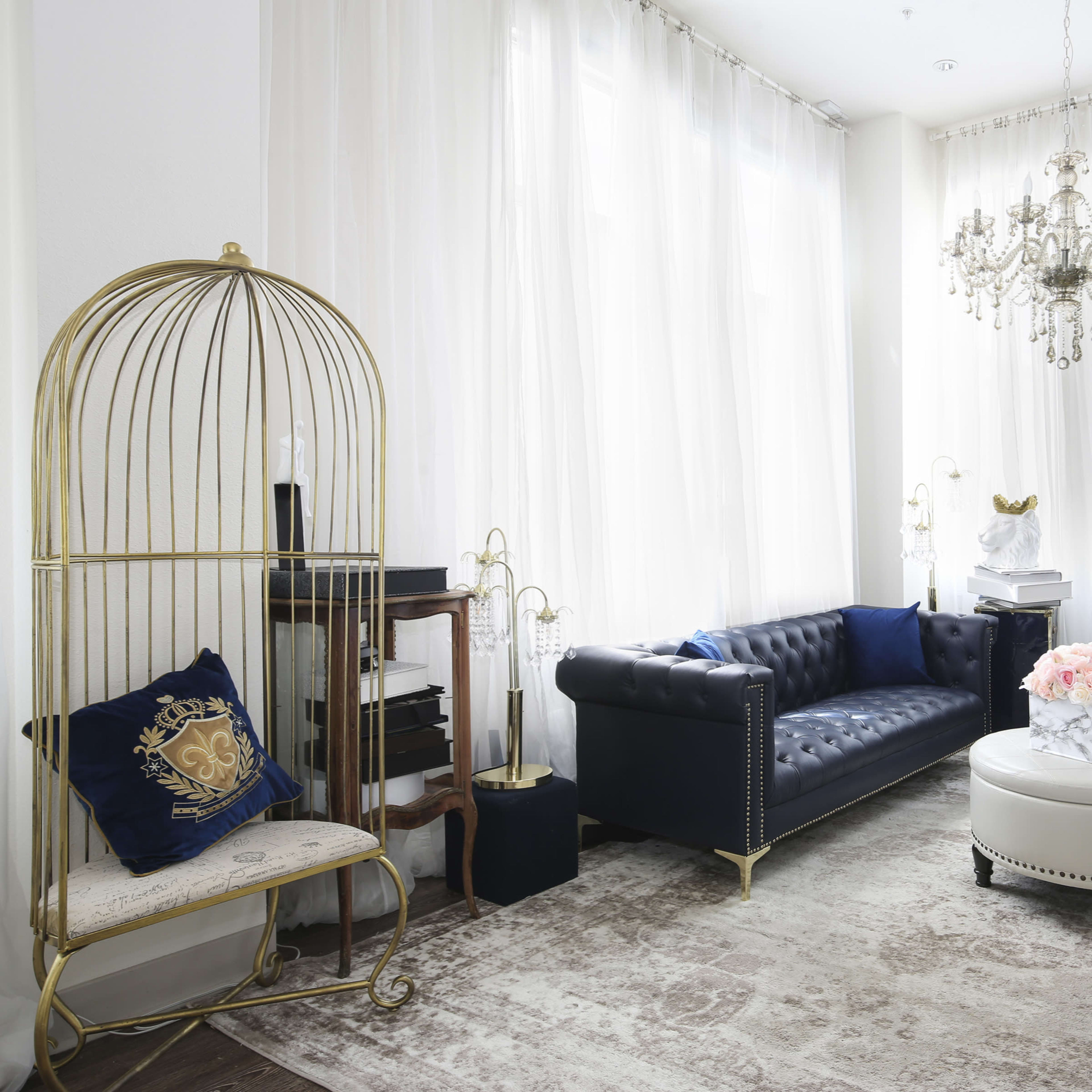 A stylish living room with two black chesterfield sofas, a round white coffee table, a decorative chandelier, and a mannequin, all set against light, sheer curtains.