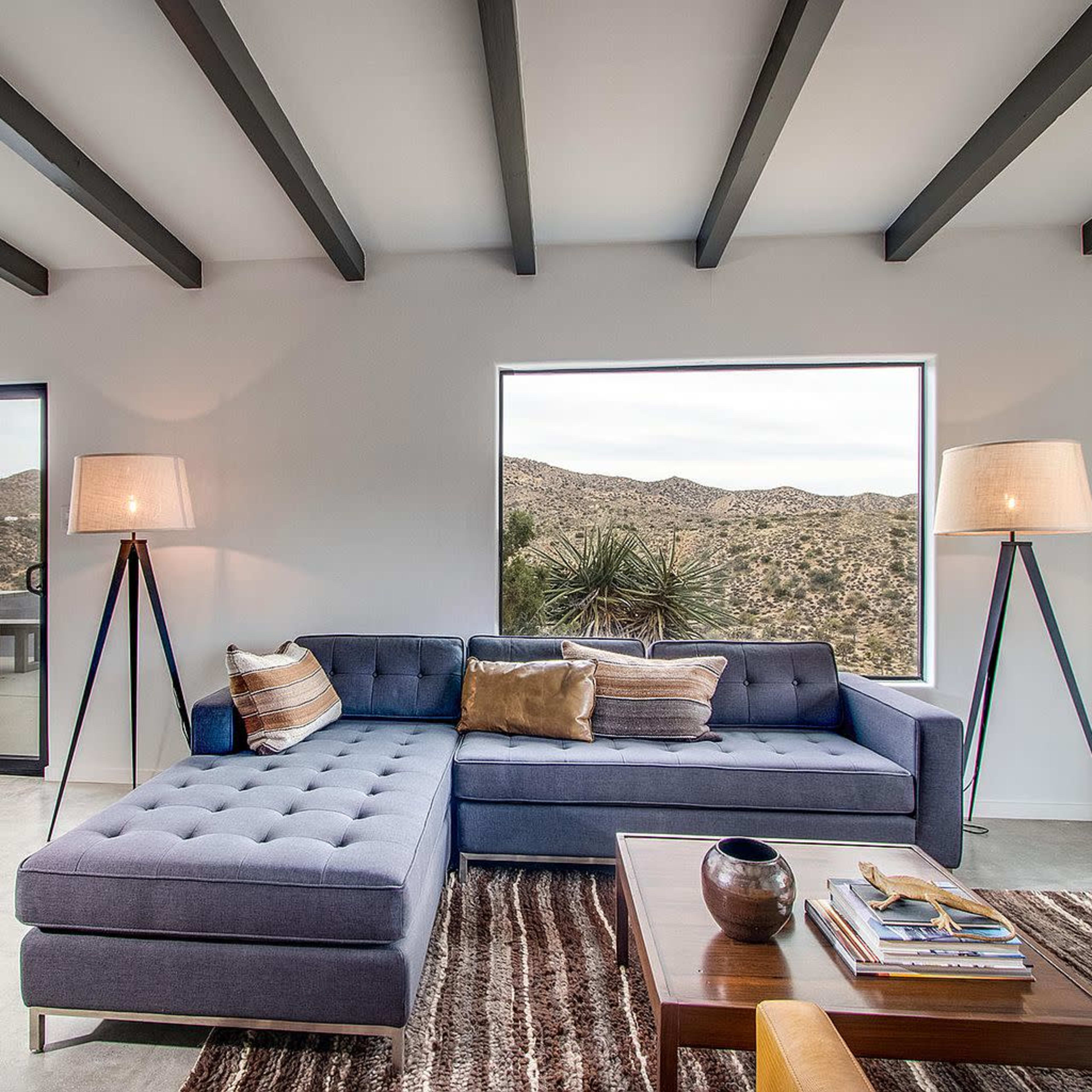 A modern living room features a blue sectional sofa, a wood coffee table, and large windows overlooking desert mountains.