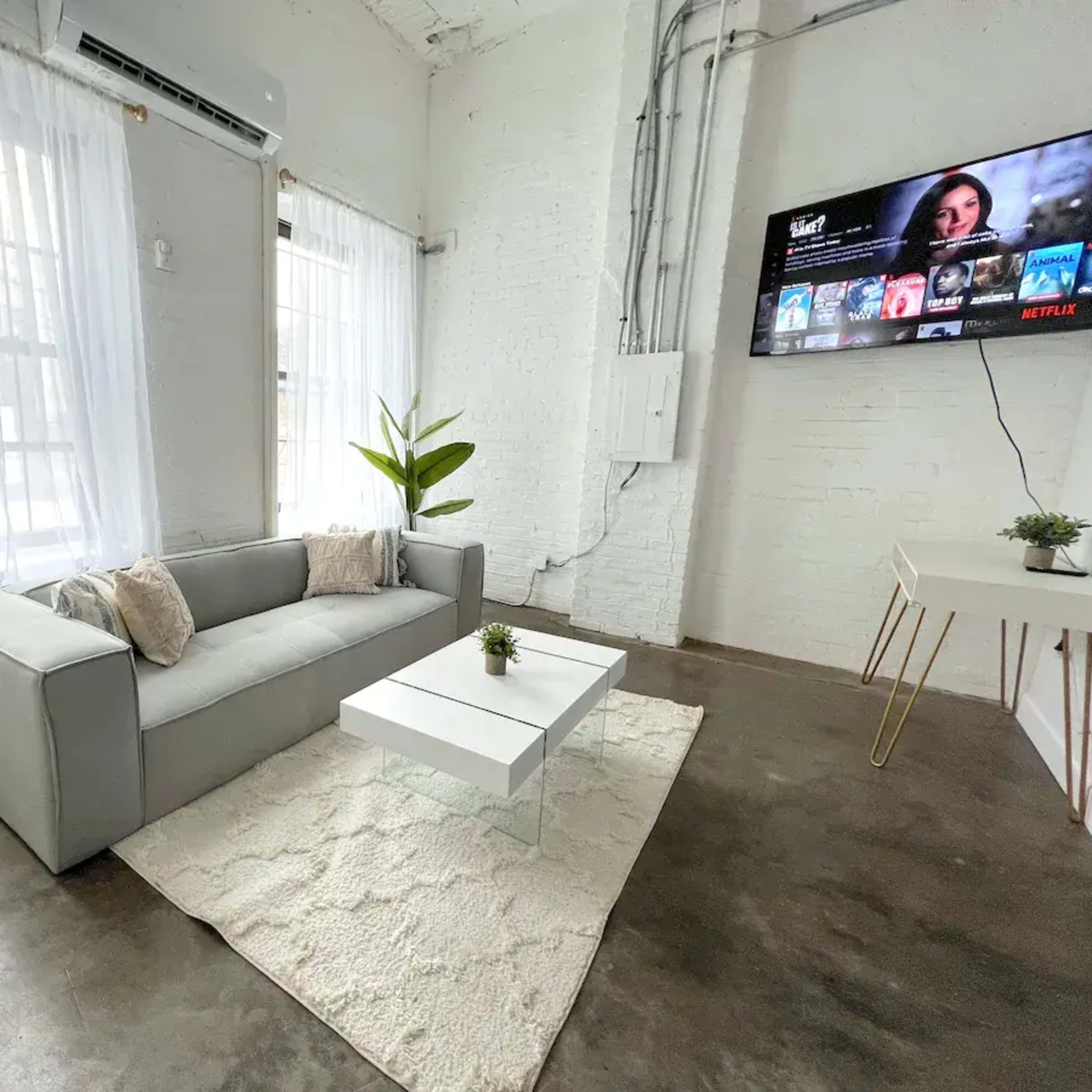 A gray sofa with decorative pillows is positioned on a light rug in a bright room featuring large windows and a wall-mounted television.