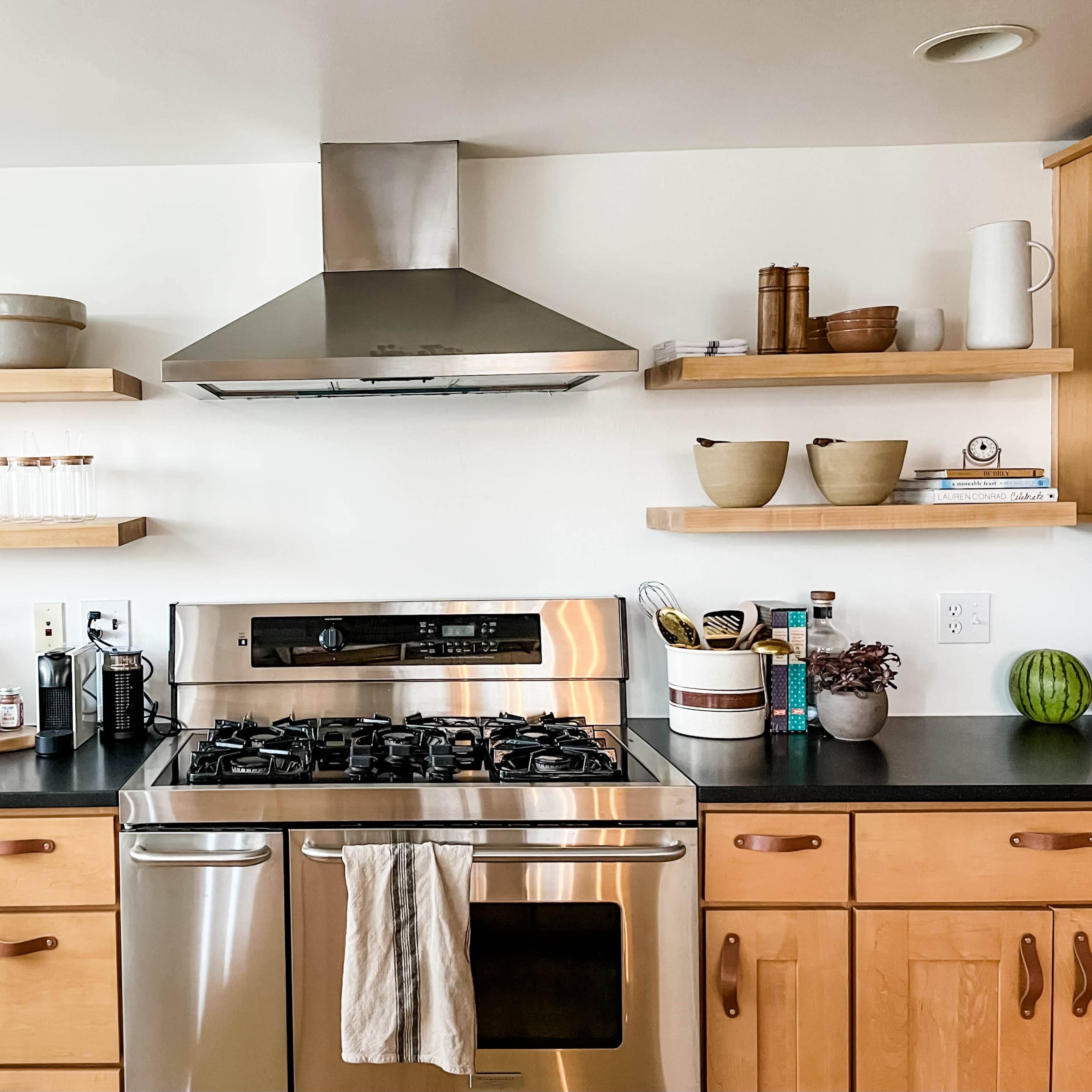 A modern kitchen with a stainless steel stove, a metallic range hood, and wooden shelves displaying various kitchen items.