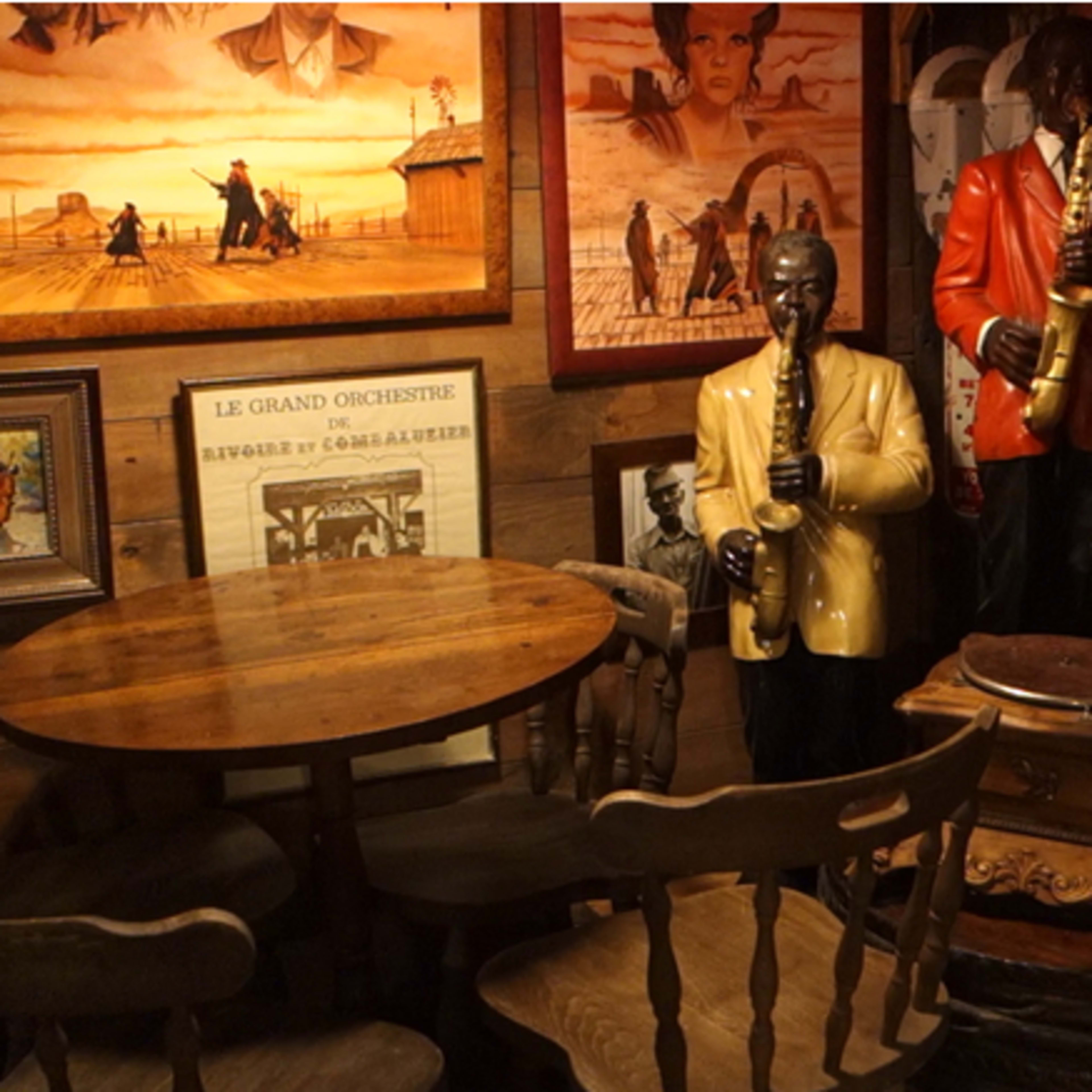 A vintage-themed room with a round wooden table surrounded by wooden chairs, and life-sized statues of musicians in colorful outfits, accompanied by framed artwork and an old-fashioned gramophone.