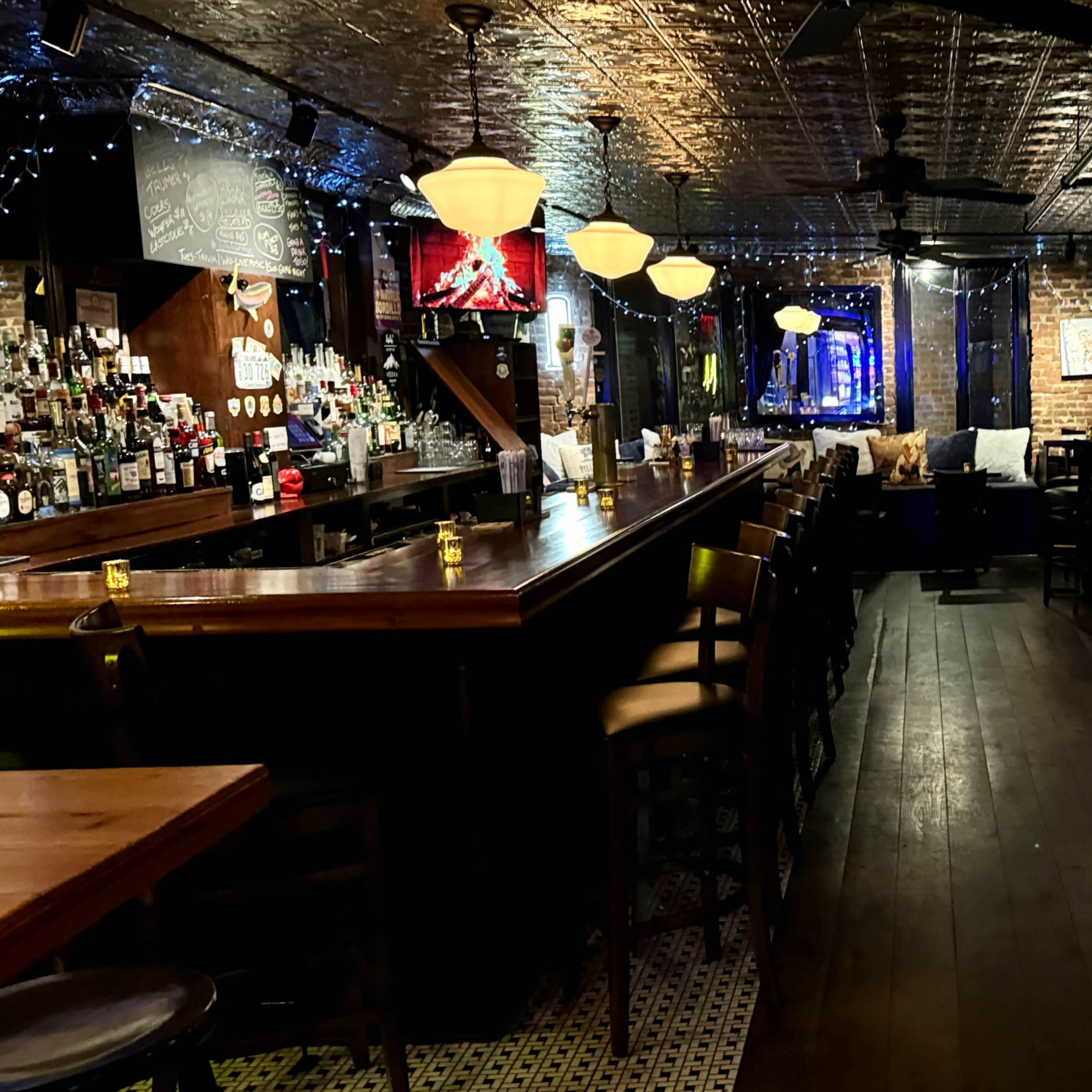 A dimly lit bar with a long wooden counter, stools, and scattered tables, adorned with string lights and artwork on the walls.