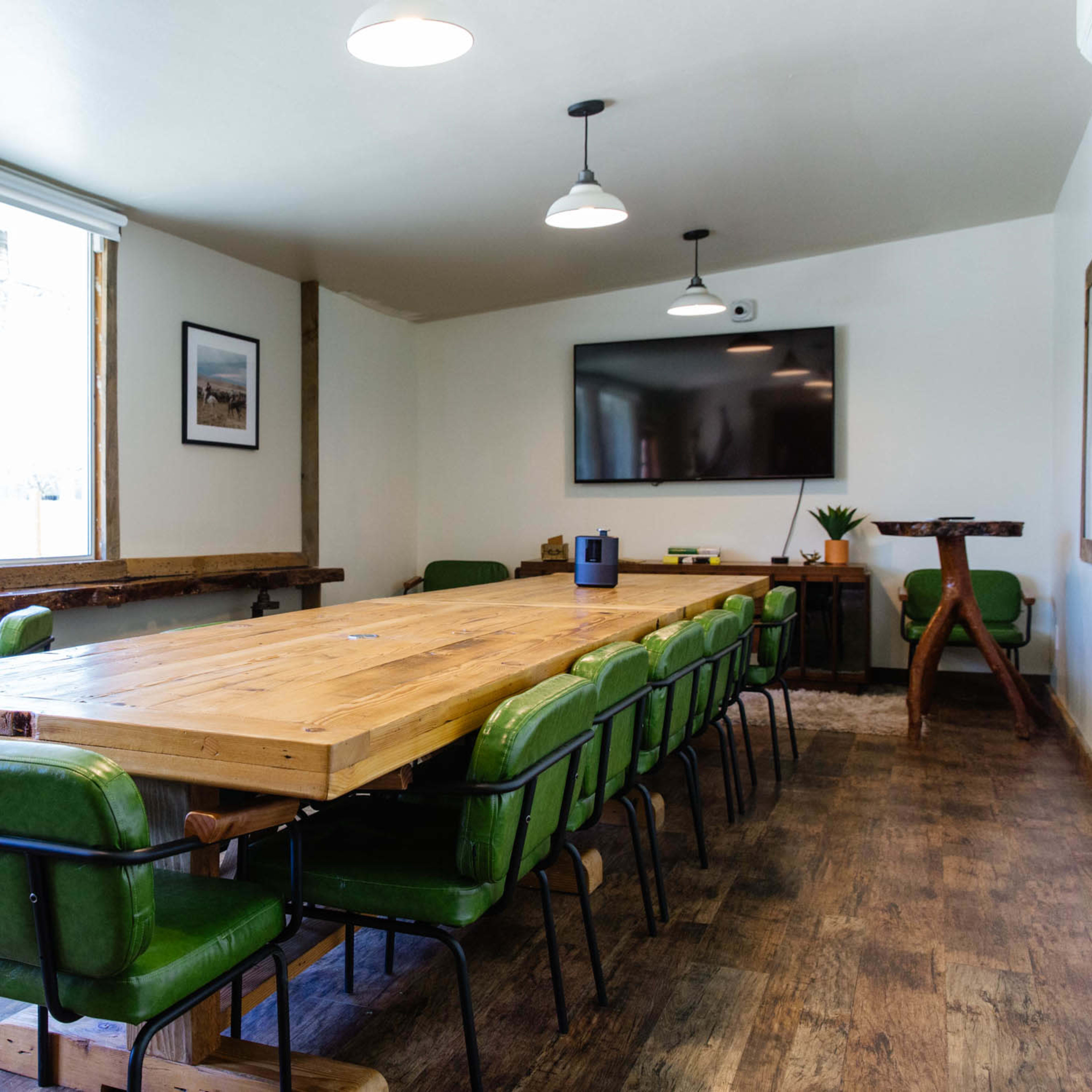 A modern conference room with a long wooden table surrounded by green chairs, large windows, and a wall-mounted television.