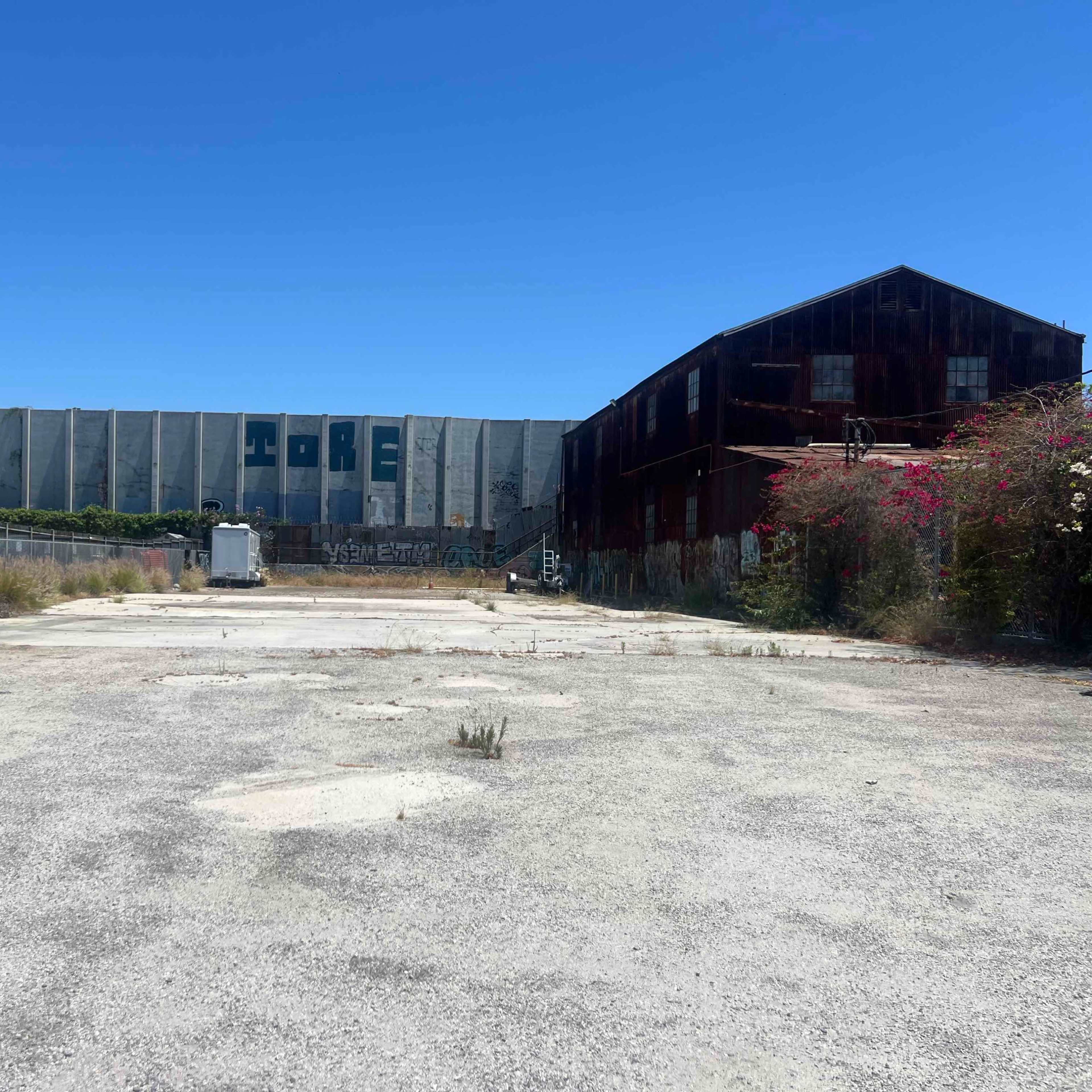 The scene shows an empty, gravel-covered lot with two large, dilapidated industrial buildings in the background under a clear blue sky.