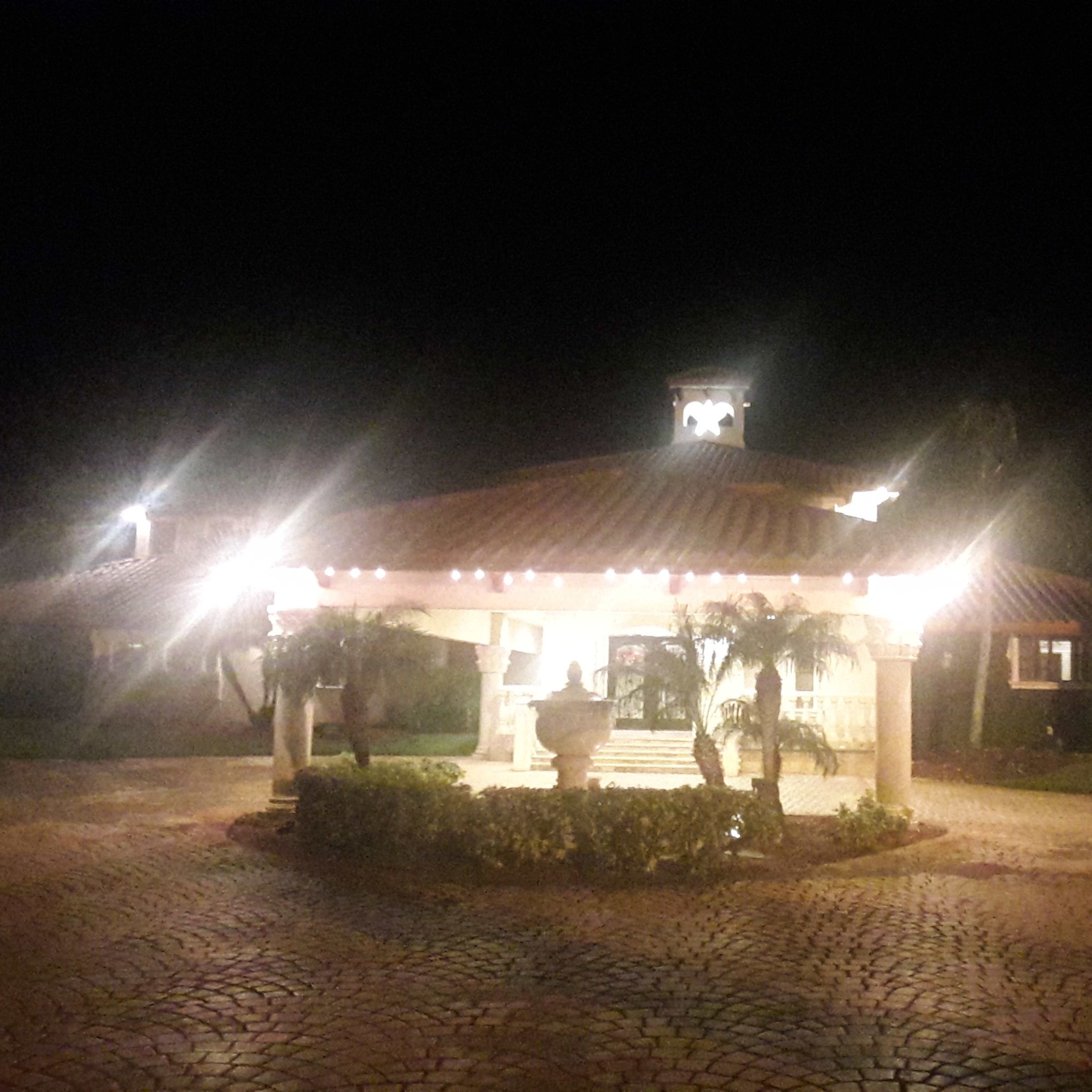 A well-lit pavilion with a tiled roof is surrounded by greenery and decorative lighting at night.