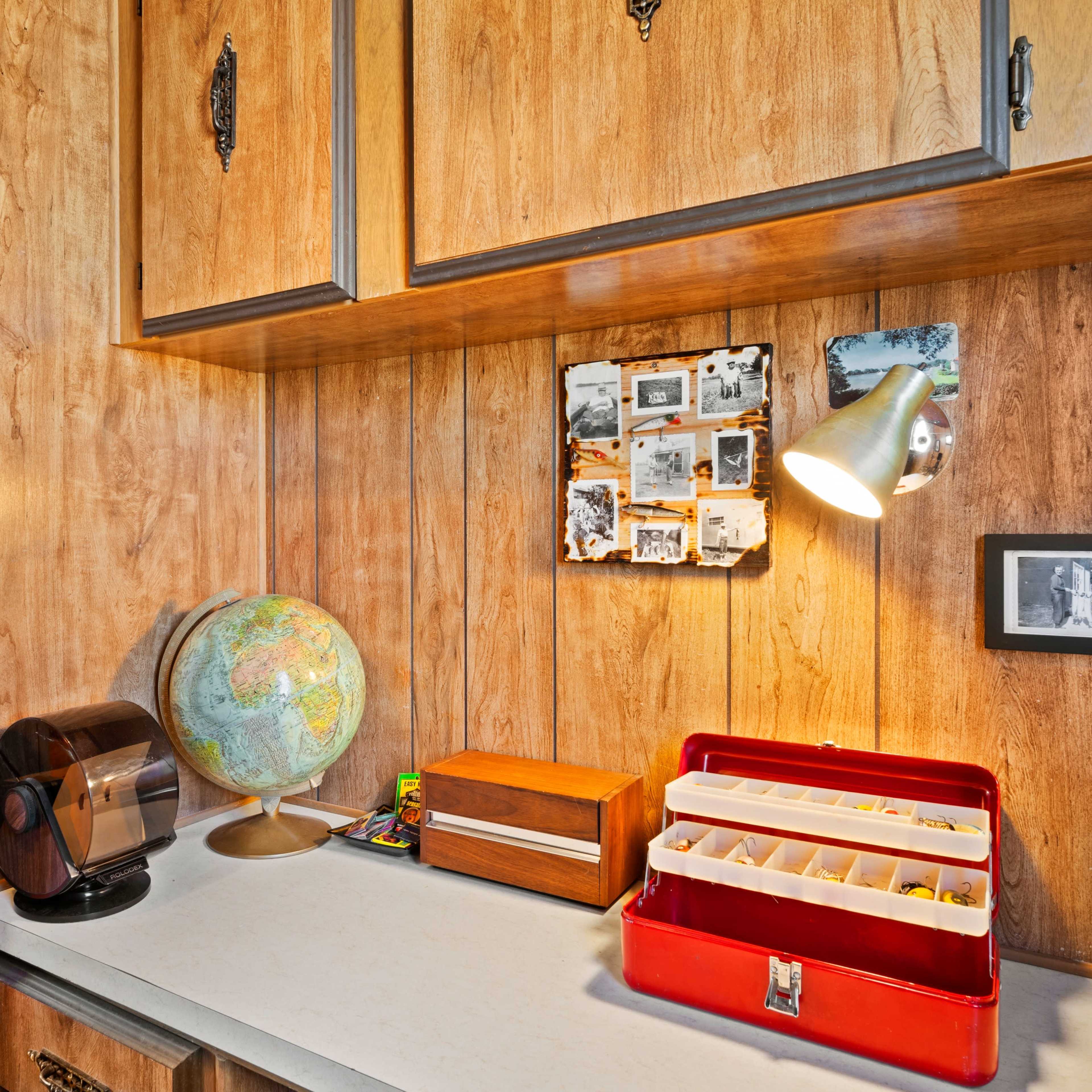 A wooden-paneled storage room features a globe, a red toolbox, and framed photographs on the wall under a mounted light.